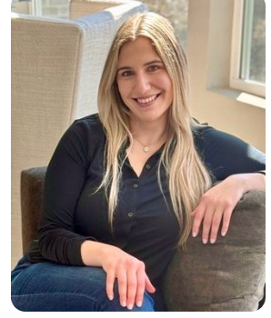 A young woman with long blonde hair smiling while sitting on a couch indoors, with a window in the background.