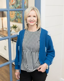 A therapist with blonde hair smiling, wearing a blue cardigan over a black-and-white checkered blouse, standing in front of a white wall with a window that has colorful decorations.