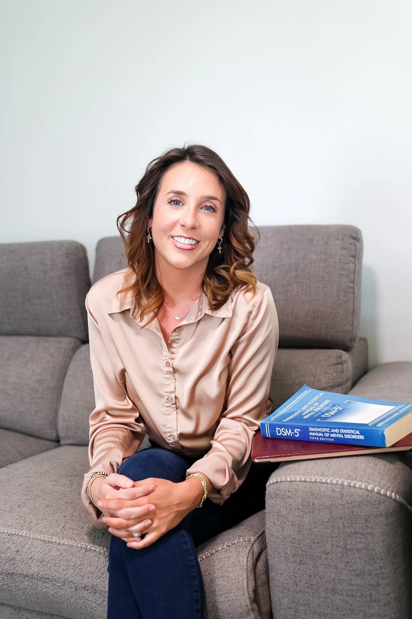A woman with shoulder-length brown hair and blue eyes, smiling while sitting on a gray couch. She is wearing a beige blouse, dark blue jeans, and jewelry. In front of her are two books, one about DSM-5 and mental disorders.
