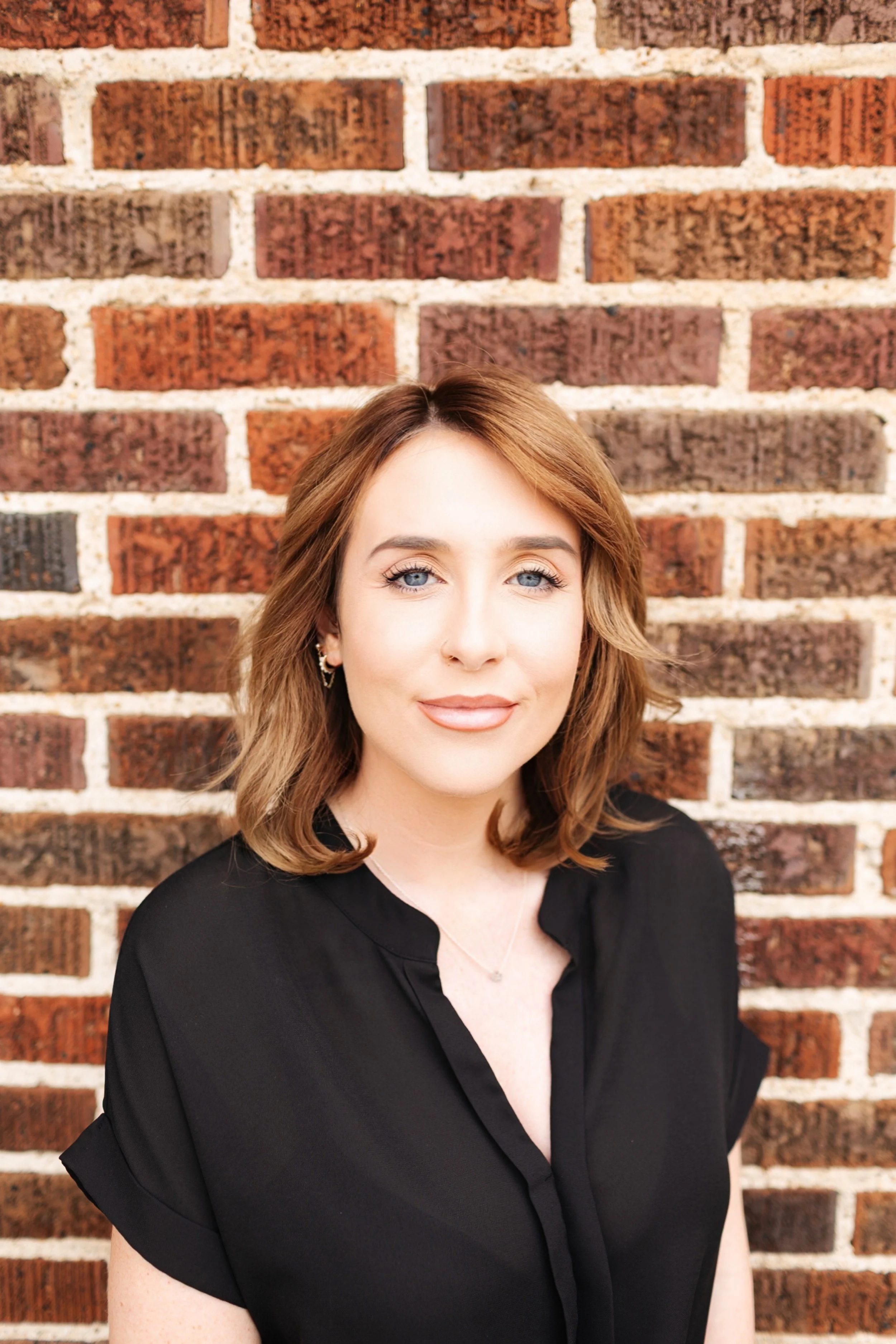 A woman with shoulder-length brown hair and blue eyes standing in front of a brick wall, wearing a black blouse with ruffled sleeves and a subtle smile.