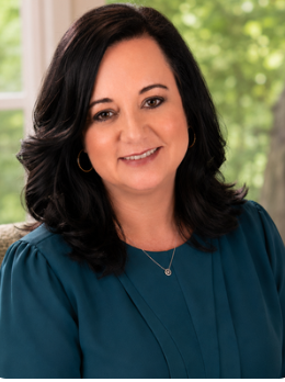 Portrait of a woman with shoulder-length black hair, wearing a black blazer and a light-colored top, smiling against a plain background.