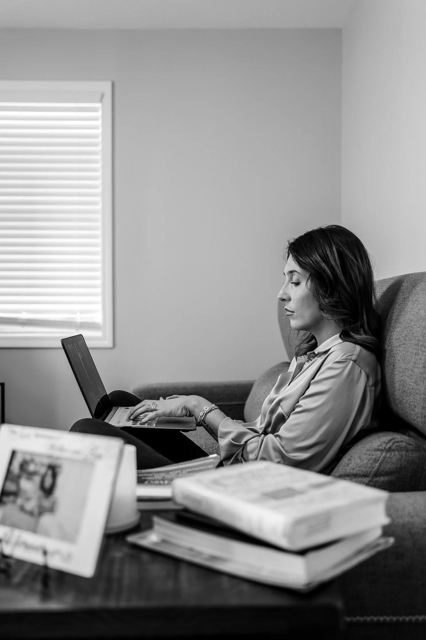 A woman with dark hair and a patterned blouse sitting on a sofa, typing on a laptop. In the foreground, there are stacks of books and magazines on a table. A window with horizontal blinds is on the wall behind her.