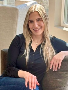 A young woman with long blonde hair smiling while sitting on a couch indoors, with a window in the background.