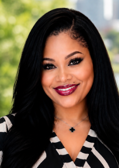 Close-up of a smiling therapist with long black hair, wearing dark makeup, a black necklace, and a black and white striped top, outdoors with blurred green background.