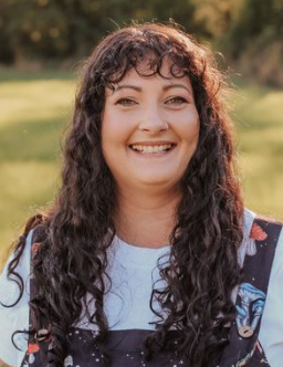 Woman with curly dark hair smiling outdoors in a grassy area