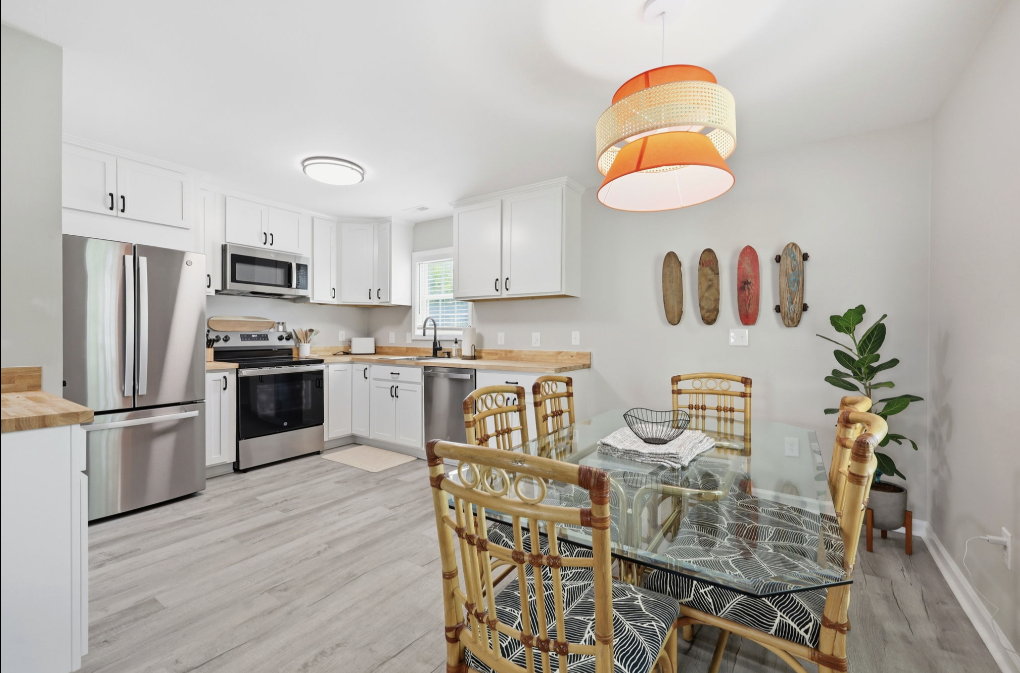 A modern kitchen with white cabinets, stainless steel appliances, a glass dining table with wicker chairs, a hanging orange and cream pendant light, wood-style flooring, wall art of skateboards, and a potted plant.