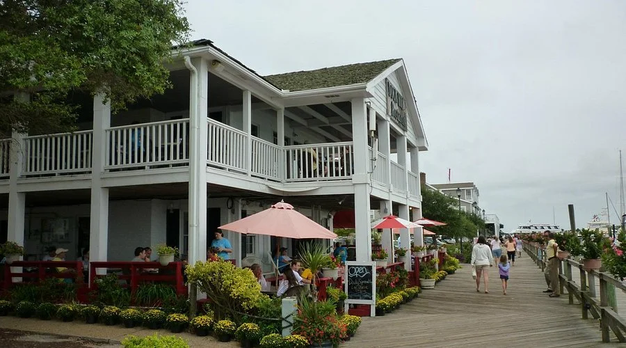 A two-story waterfront restaurant with outdoor seating, pink umbrellas, and a board indicating it is open, located on a wooden pier with people walking along.