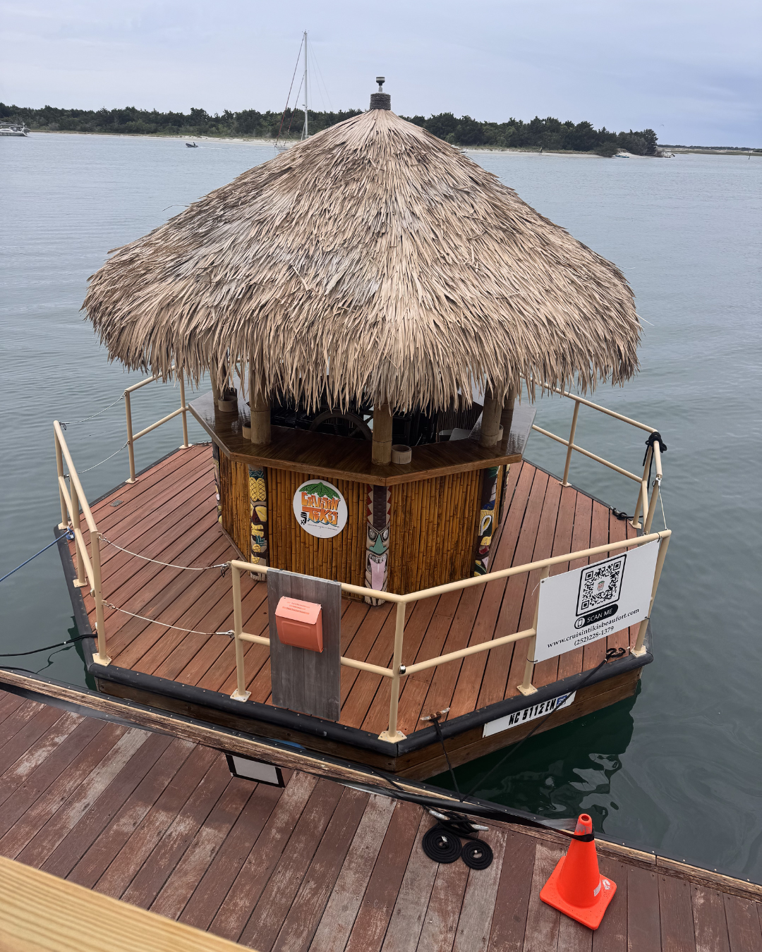 A tiki hut with a thatched roof on a floating platform in the water, surrounded by a beige railing, with a QR code and a logo on the front.