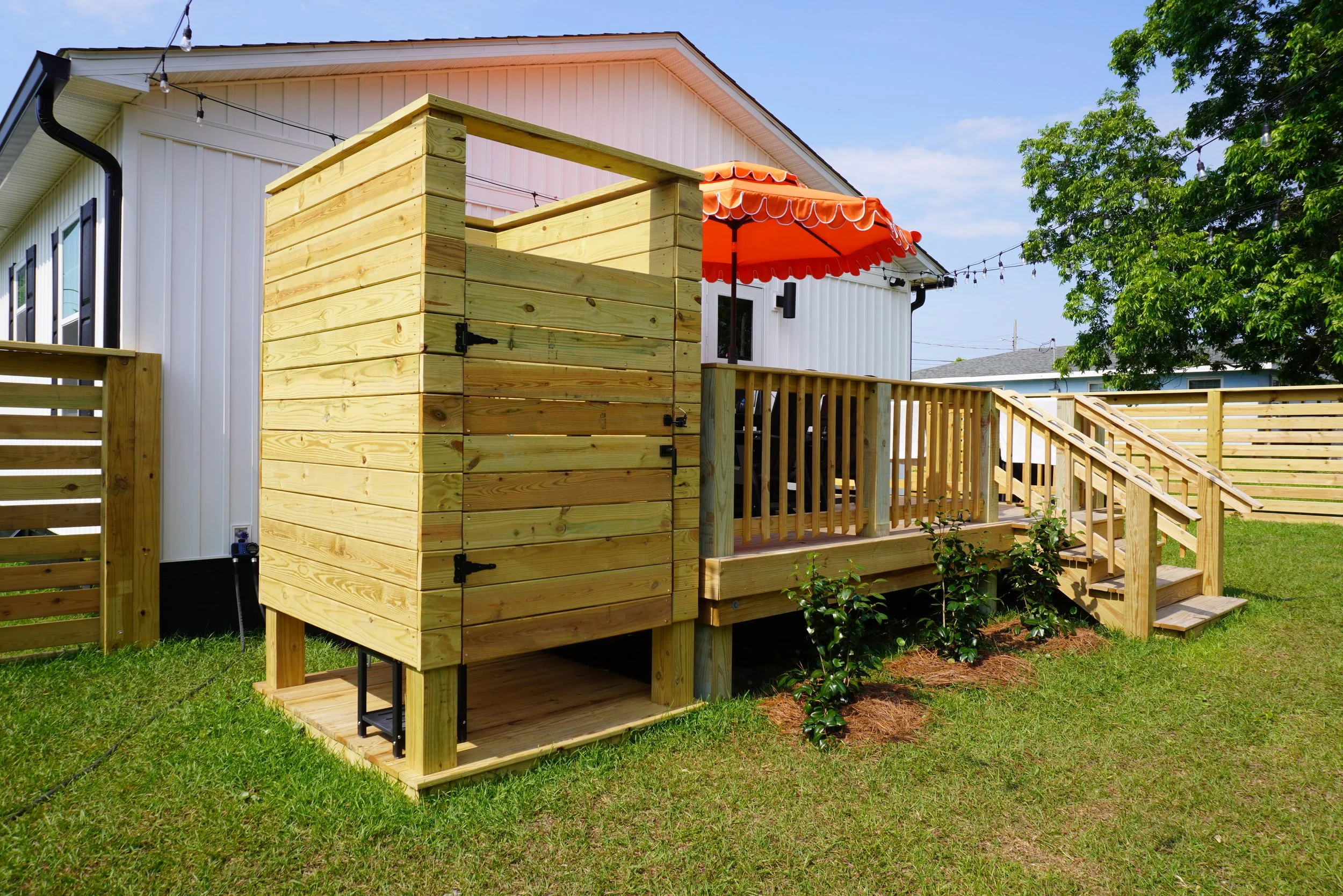 A backyard scene featuring a small elevated deck with a staircase, surrounded by a wooden fence. There's a house in the background with a white exterior and a red-and-white striped patio umbrella. String lights are hanging across the yard, and a large green tree is visible on the right.