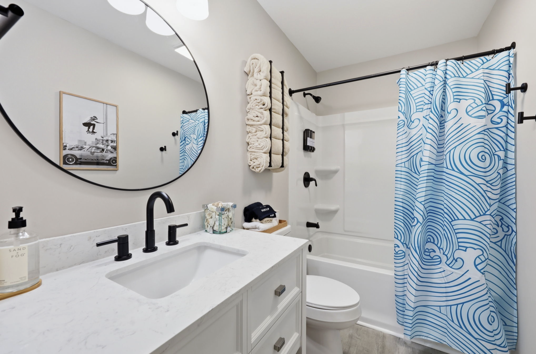 A modern bathroom with a white marble vanity, black fixtures, a large round mirror, a white bathtub with black fixtures and a blue and white patterned shower curtain, and a wall-mounted towel rack with rolled beige towels.