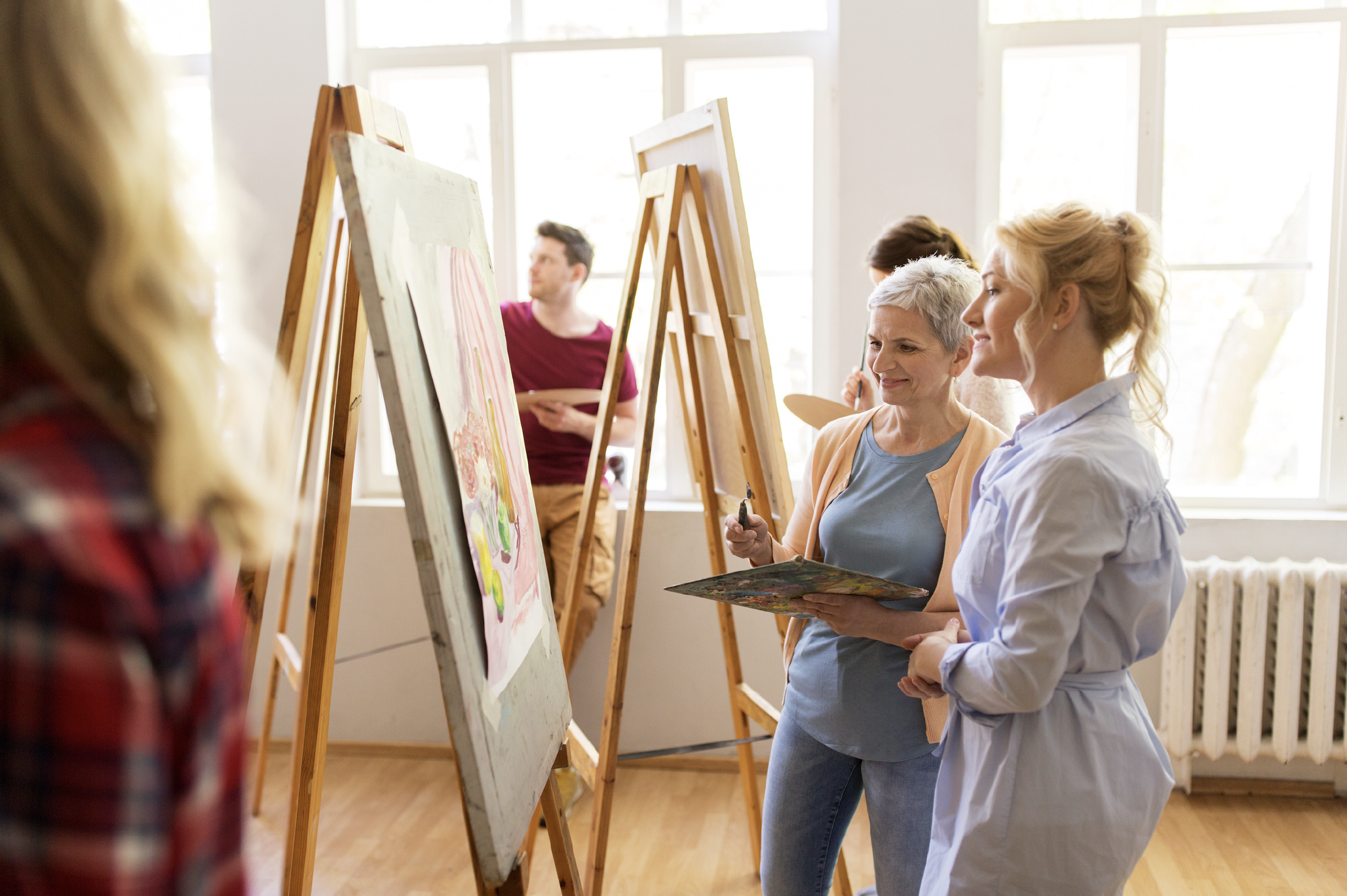 Group of people participating in a painting class, standing at easels, with large windows in a well-lit room.