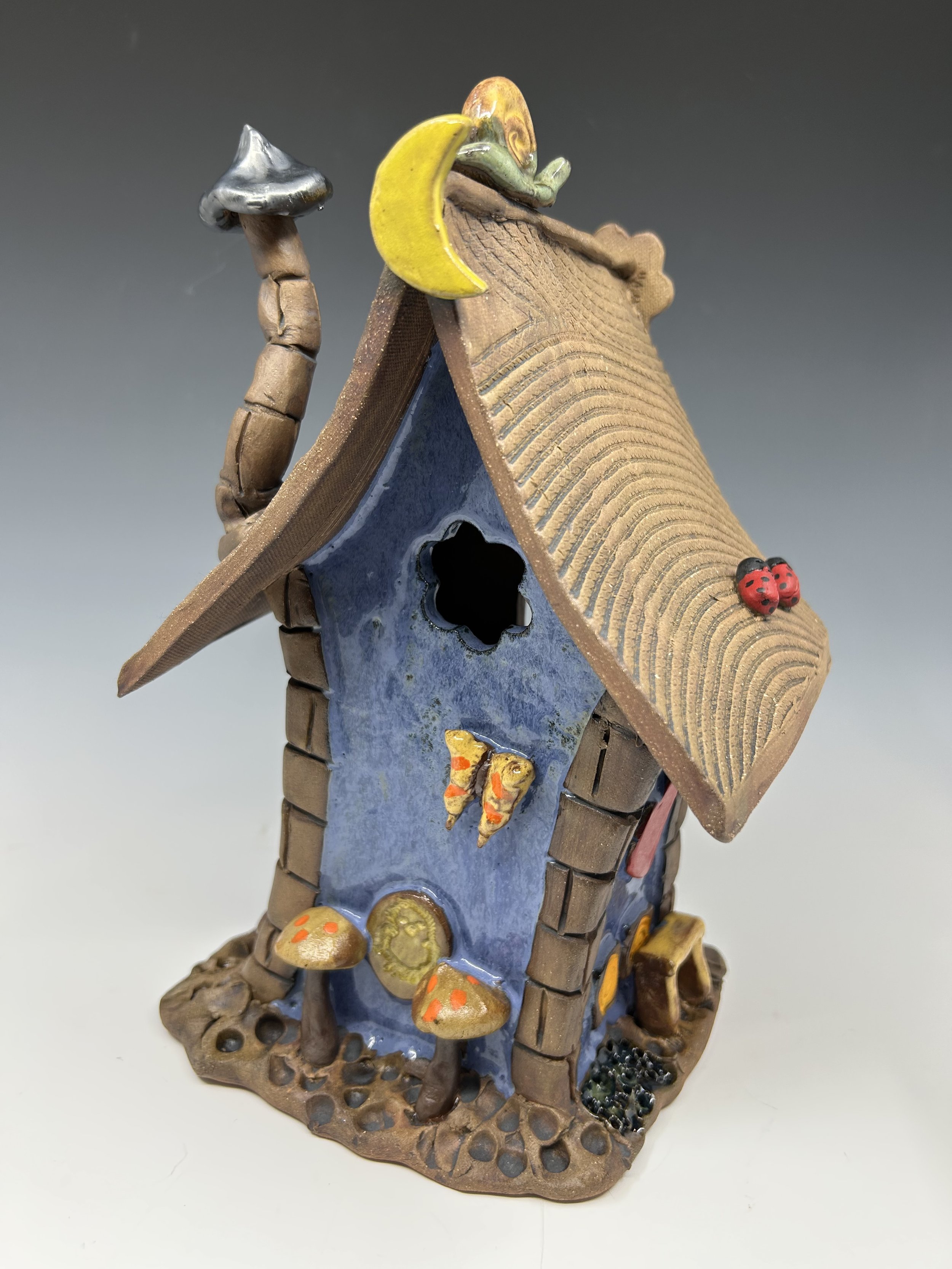 A decorative ceramic house with a blue front, brown roof, and small door, featuring mushrooms, ladybugs, a butterfly, and a snail as embellishments.