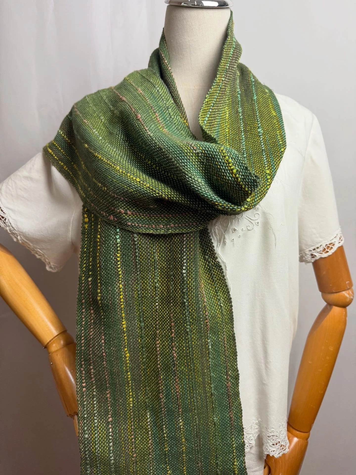 A mannequin dressed in a white embroidered dress with short sleeves and lace trim, accessorized with a green multicolored woven scarf draped around the neck and hanging down the front.