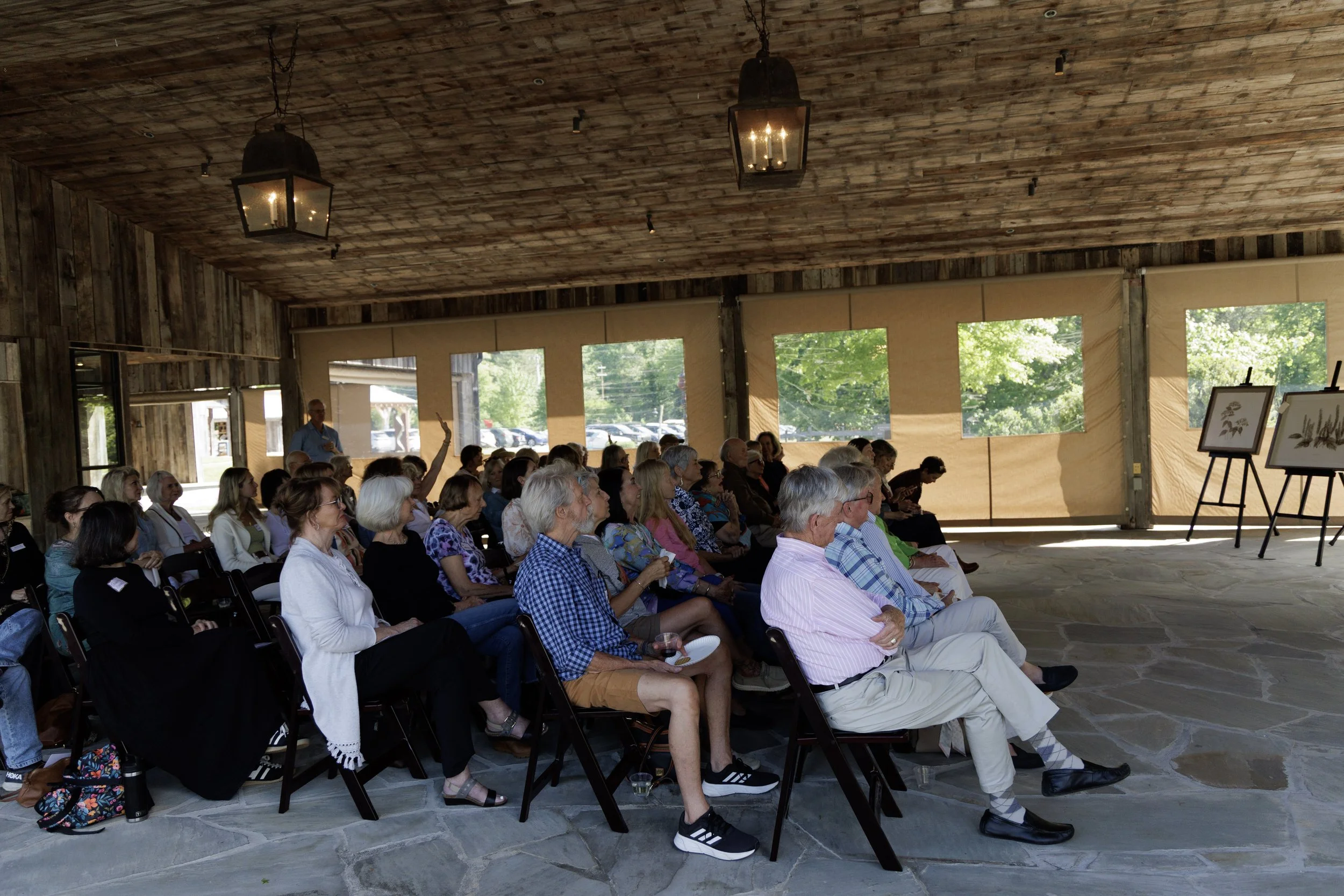 An indoor event space with wooden walls and ceiling, filled with a seated audience listening to a speaker. Some audience members have raised their hands. There are large windows with natural light and framed artwork on display.