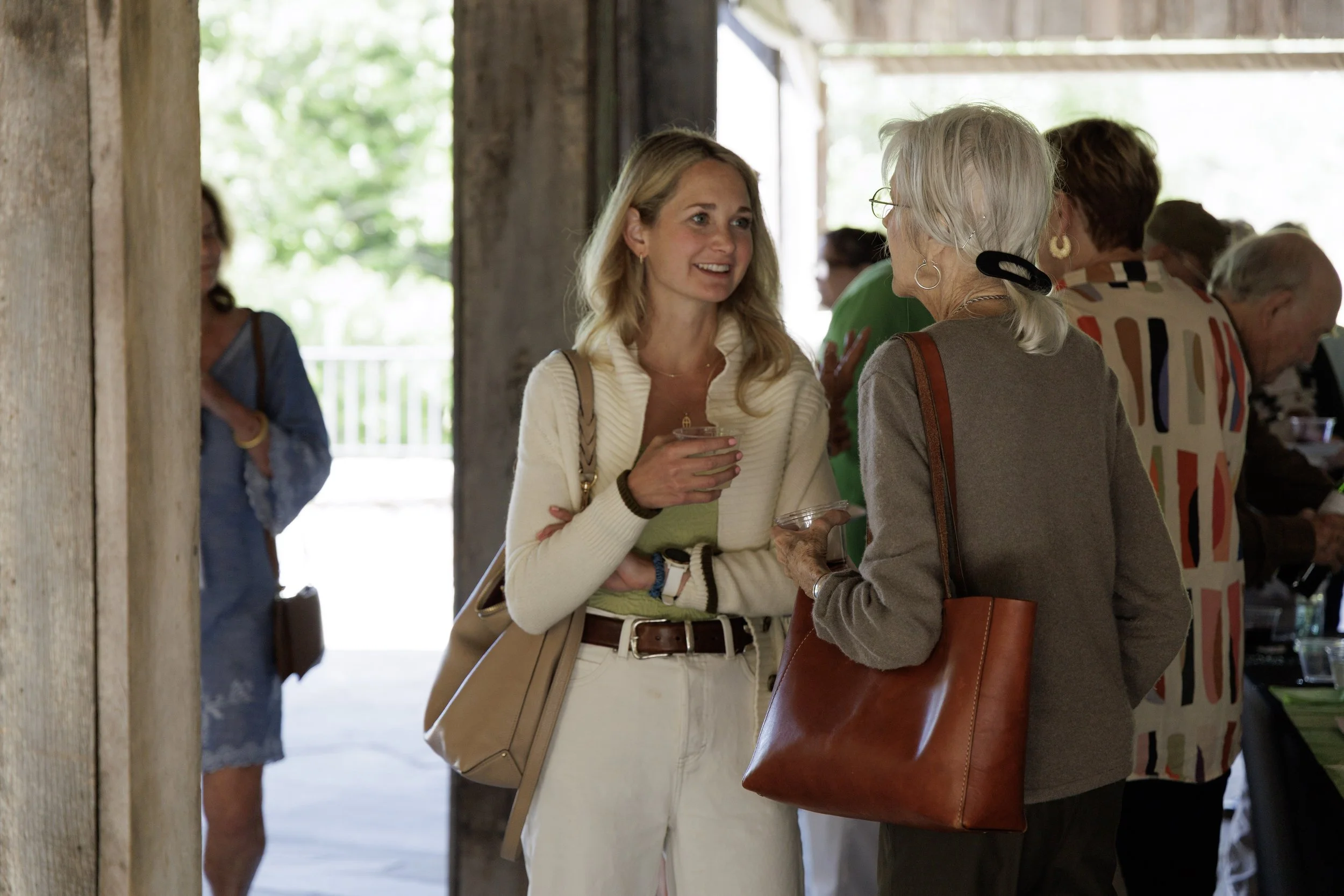 Two women are engaged in conversation at an indoor social gathering, with several other people in the background.