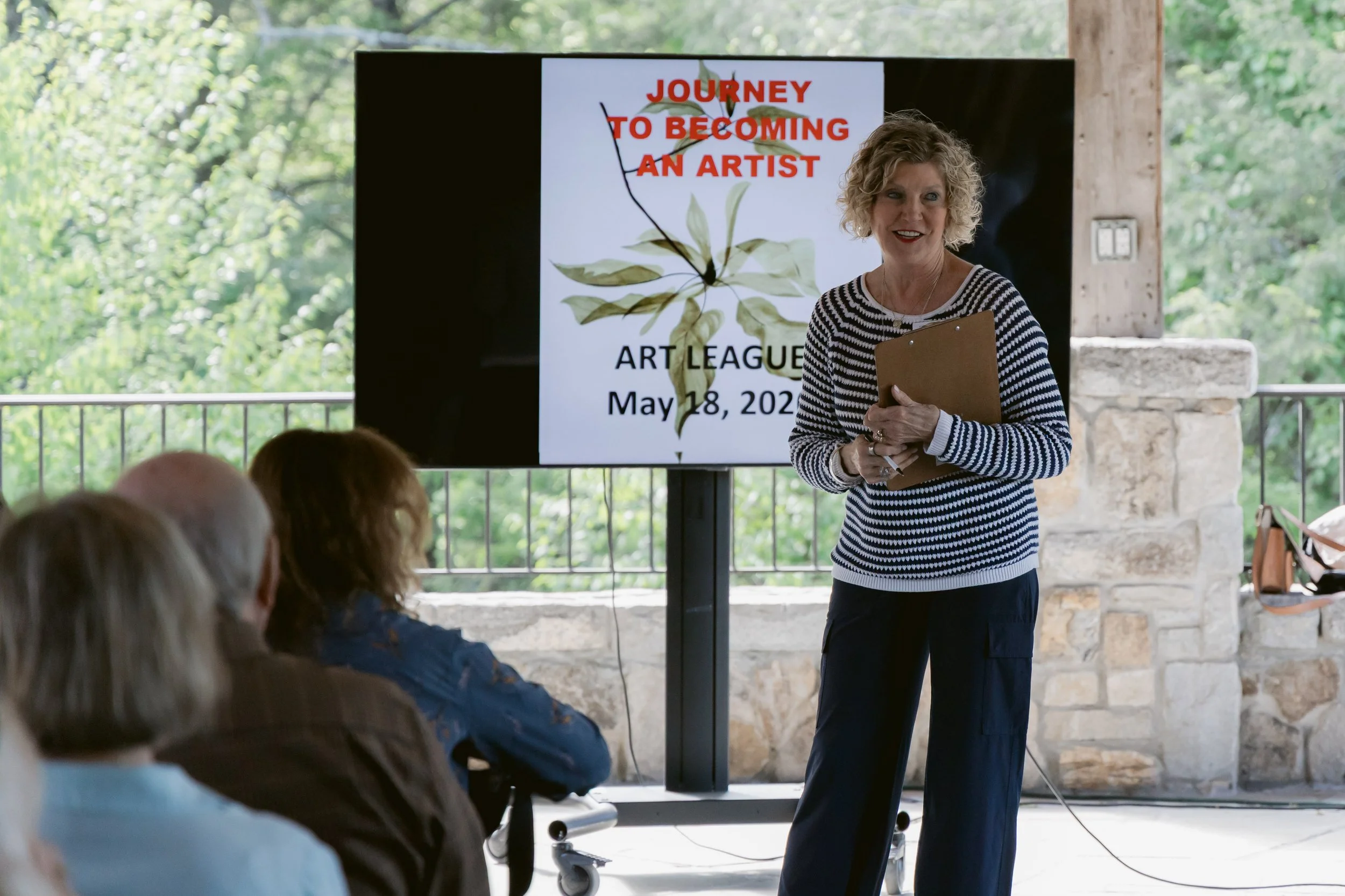 A woman giving a presentation in an outdoor setting with a large screen behind her displaying the text 'Journey to Becoming an Artist' and the date May 18, 2023. Attendees are seated and listening.