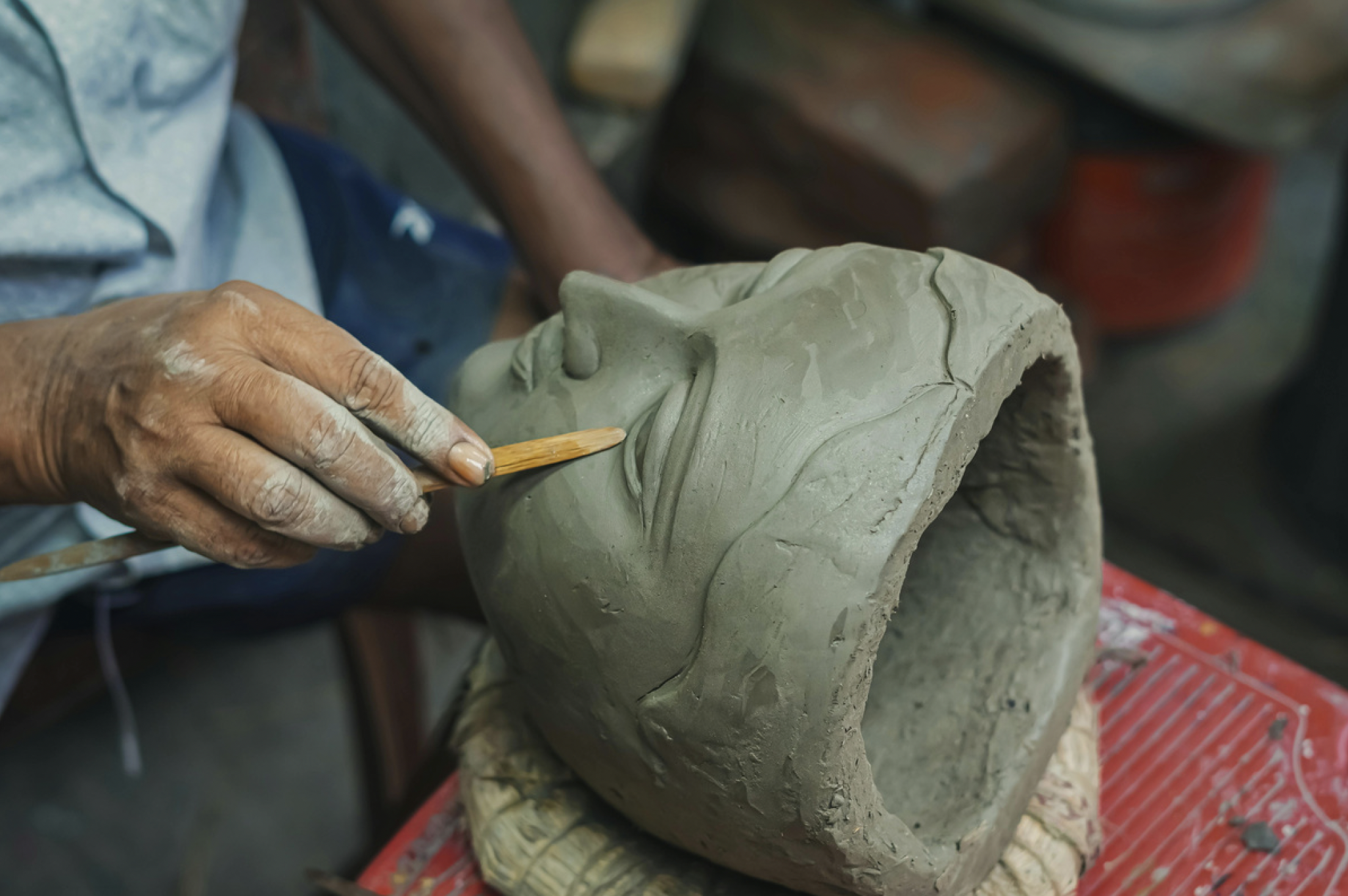 An artisan sculpting a detailed face on a clay bust, using a small sculpting tool, with a workbench and tools in the background.
