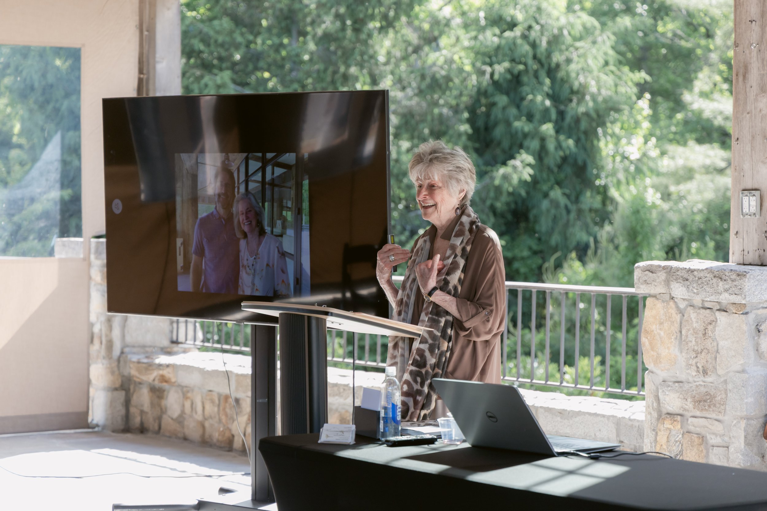 An elderly woman with gray hair smiling and talking during a video call on a large TV screen. She is standing in a bright room with large windows and green trees outside. A laptop, water bottles, and papers are on the table in front of her.