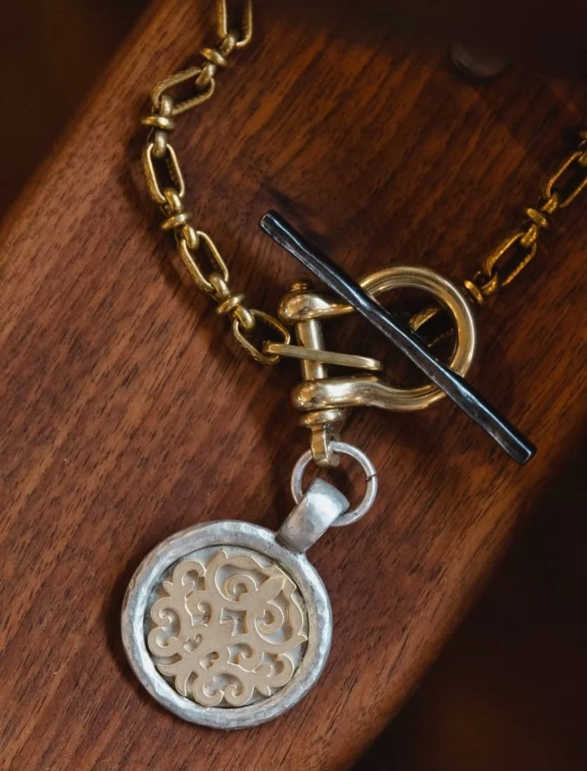 A close-up of a gold chain necklace with a lock clasp, a black toggle bar, and a decorative circular pendant with intricate cut-out patterns resting on a wooden surface.