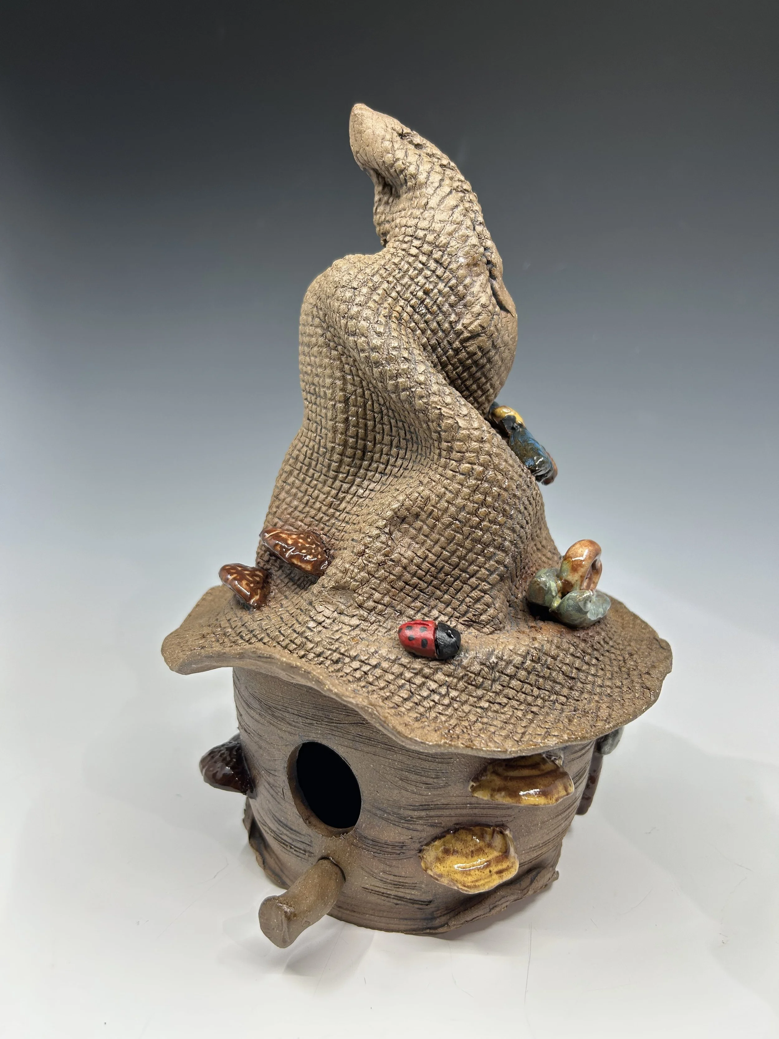 A decorative ceramic birdhouse with a textured roof, decorated with small colorful ceramic insects including ladybugs and butterflies, set against a neutral gradient background.