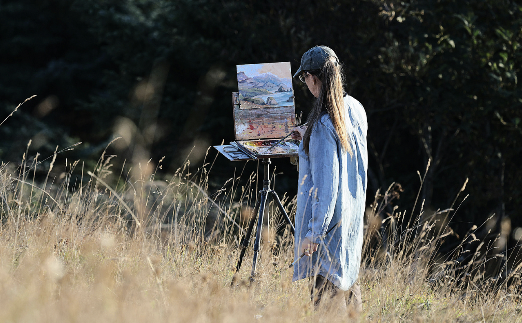 A woman painting outdoors in a grassy field with trees in the background, wearing a denim jacket, cap, and glasses, using an easel and palette.
