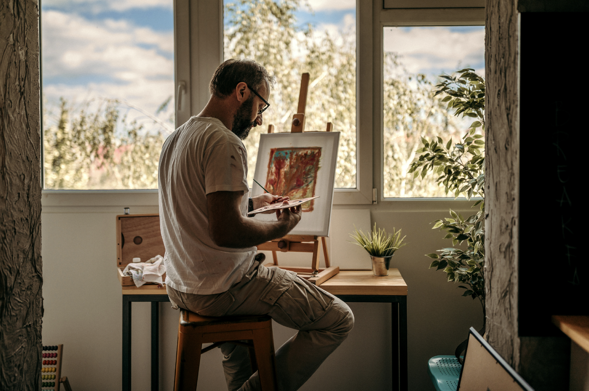 A man with glasses and a beard sitting on a stool, holding a palette and a brush, working on an abstract painting on an easel in a bright room with large windows, indoor plants, and natural light.