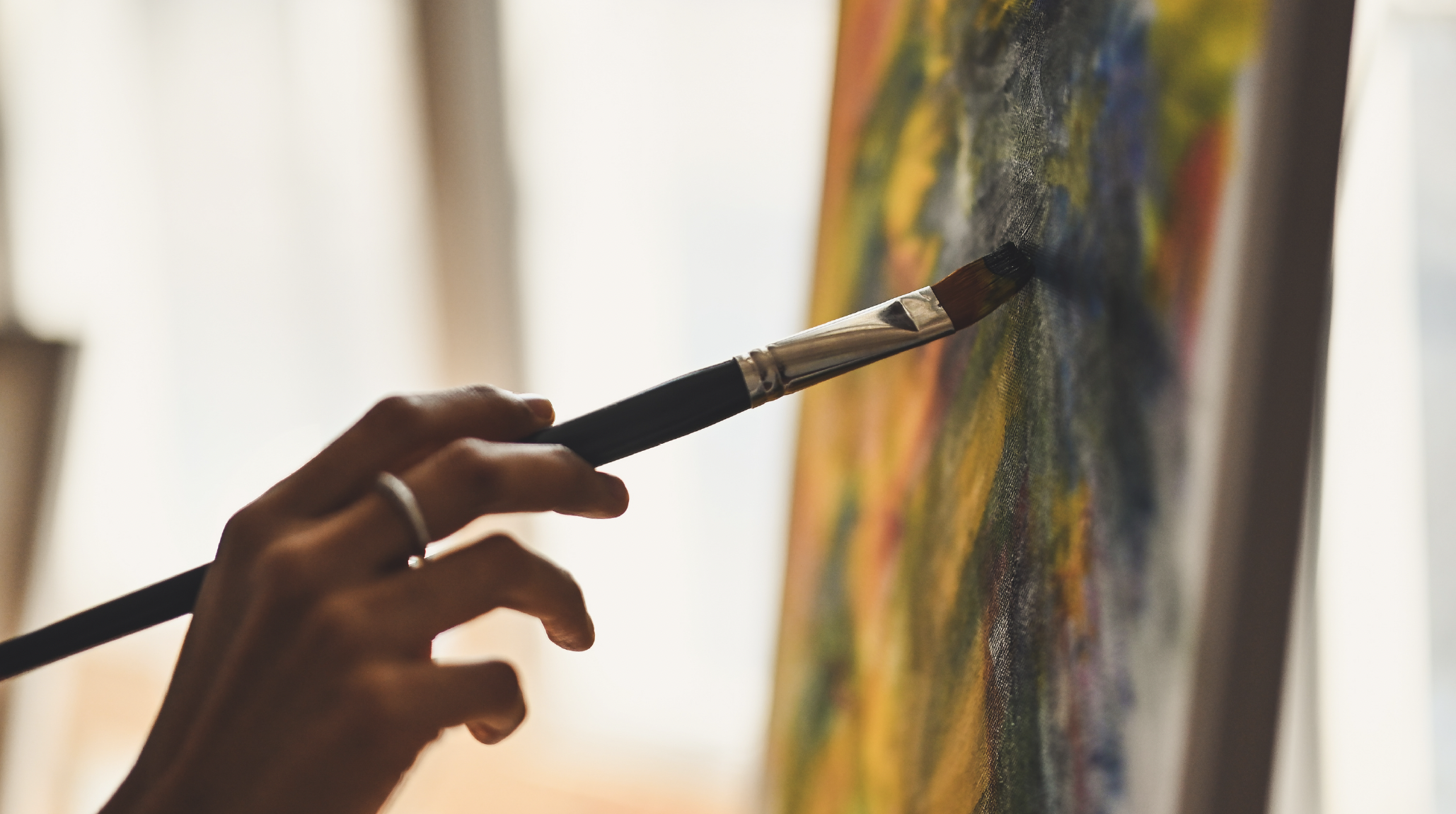 A person's hand holding a paintbrush and painting on a colorful canvas.