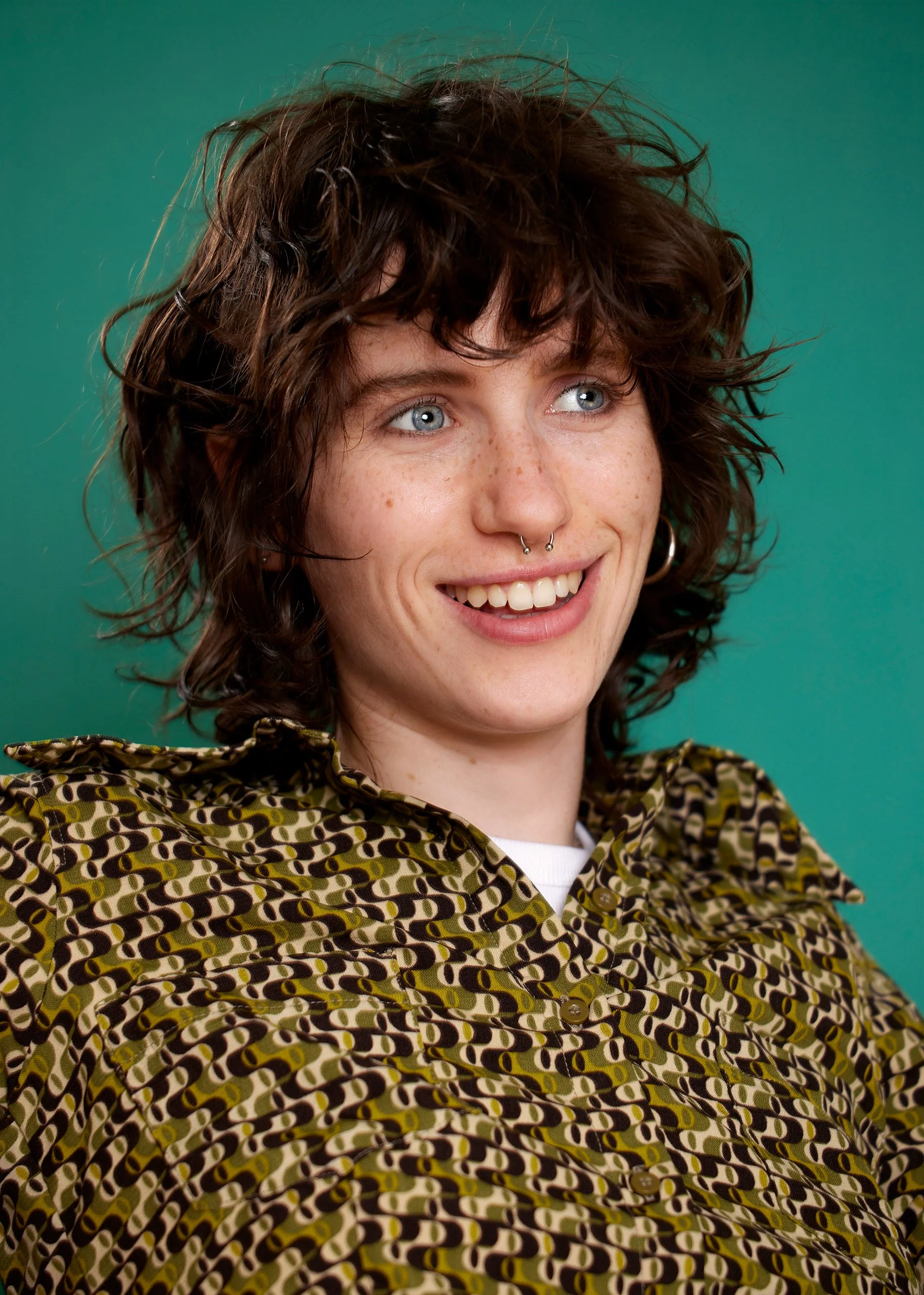 Close-up of a young woman with curly brown hair, freckles, blue eyes, and septum piercing, smiling and wearing a patterned shirt against a green background.