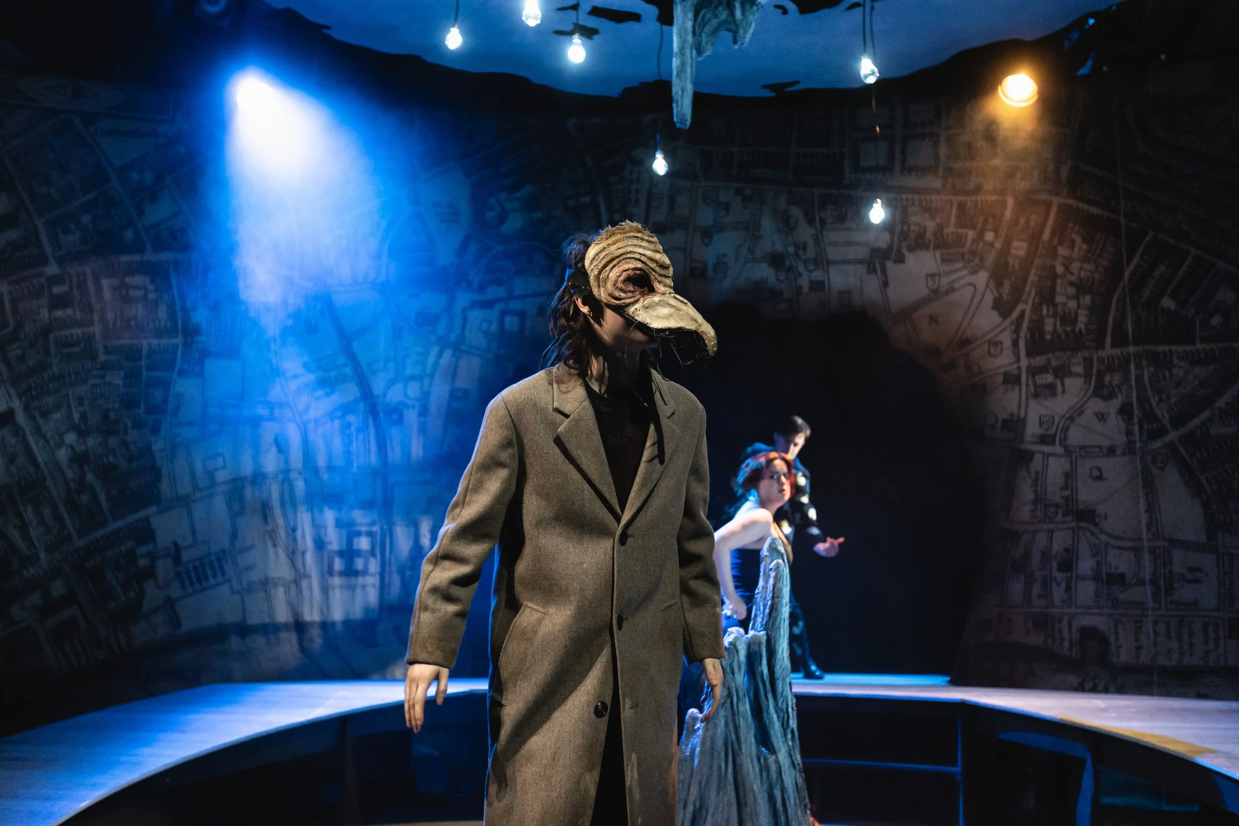 A person wearing a bird beak mask and beige coat walking in a dimly lit room with blue and orange lighting, background features other people and a map on the wall.