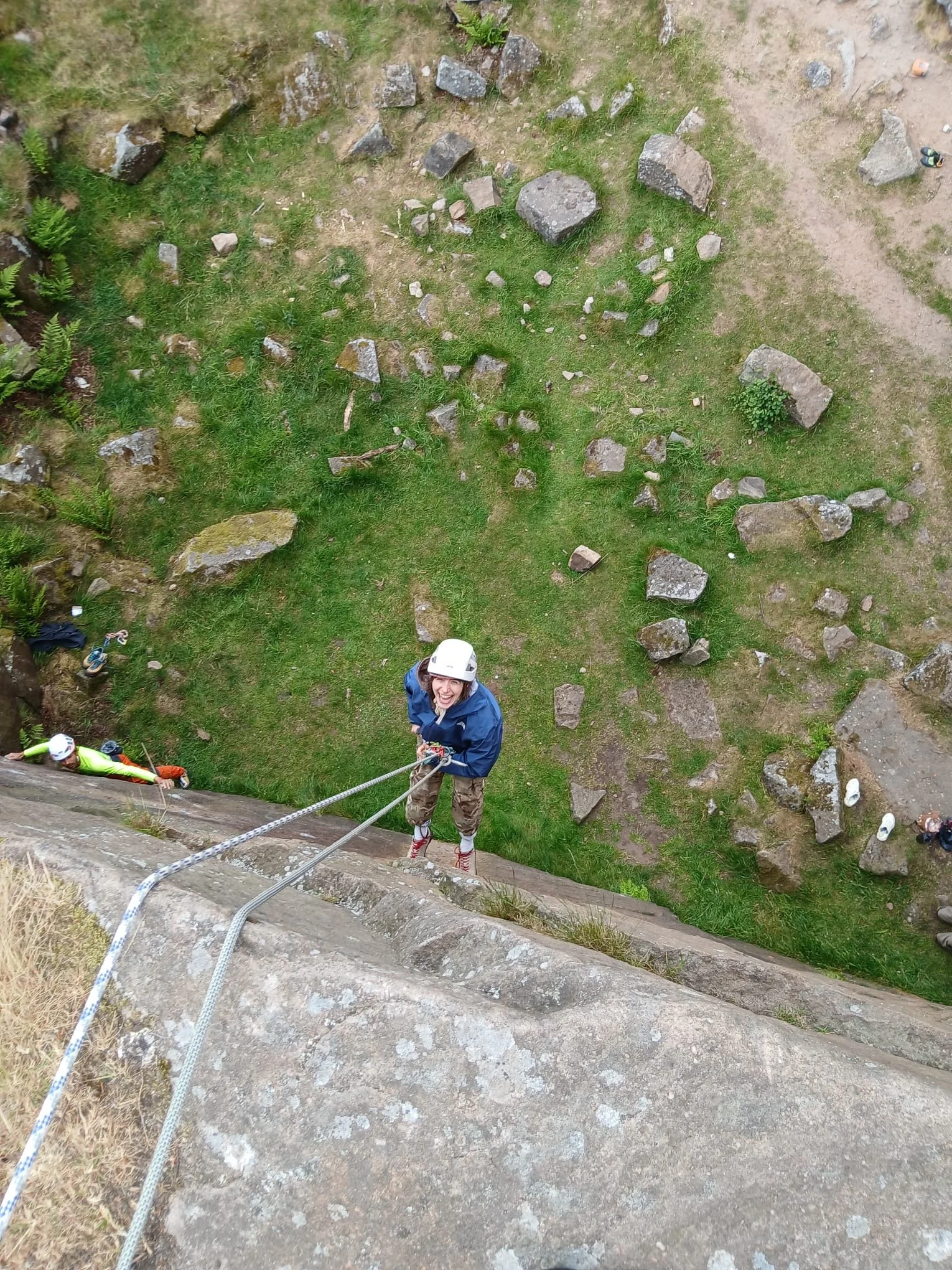A woman is rock climbing on an outdoor rock face and is secured by a harness and safety rope, looking up and smiling.