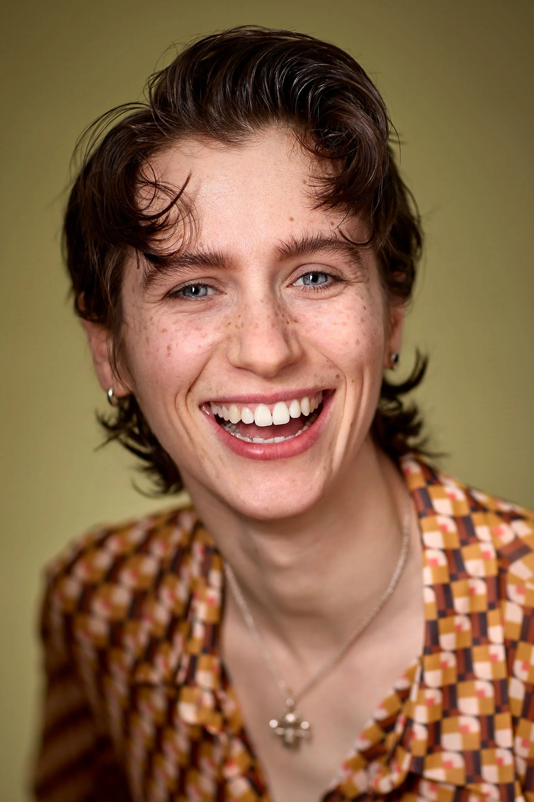 Close-up of a smiling young woman with short, wavy brown hair, blue eyes, freckles. Looks bright, friendly and welcoming.