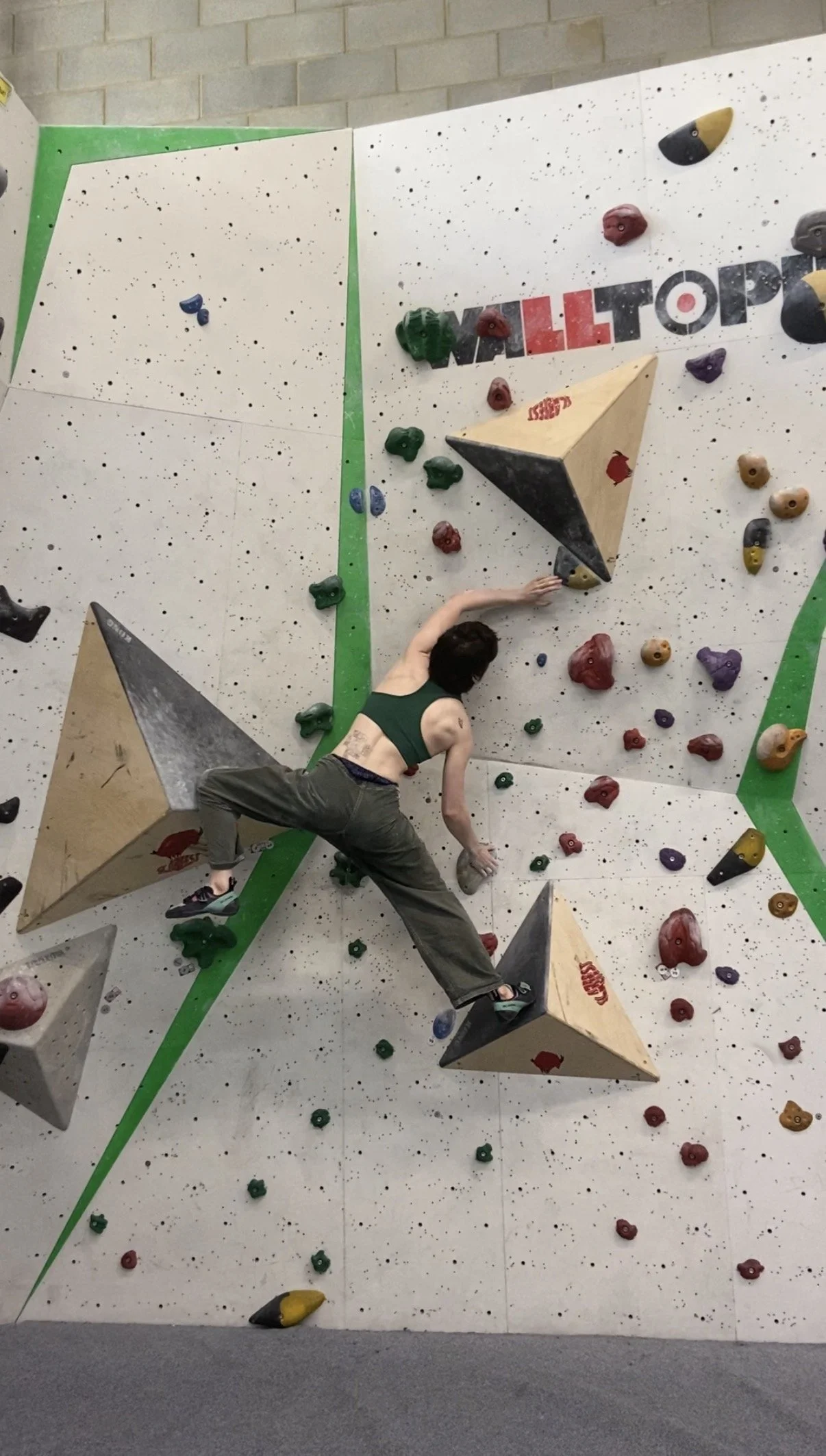 A woman is climbing an indoor bouldering wall with various colorful holds, some large and geometric, in a gym with gray walls and floor. She has strong muscles and is enjoying exercising.