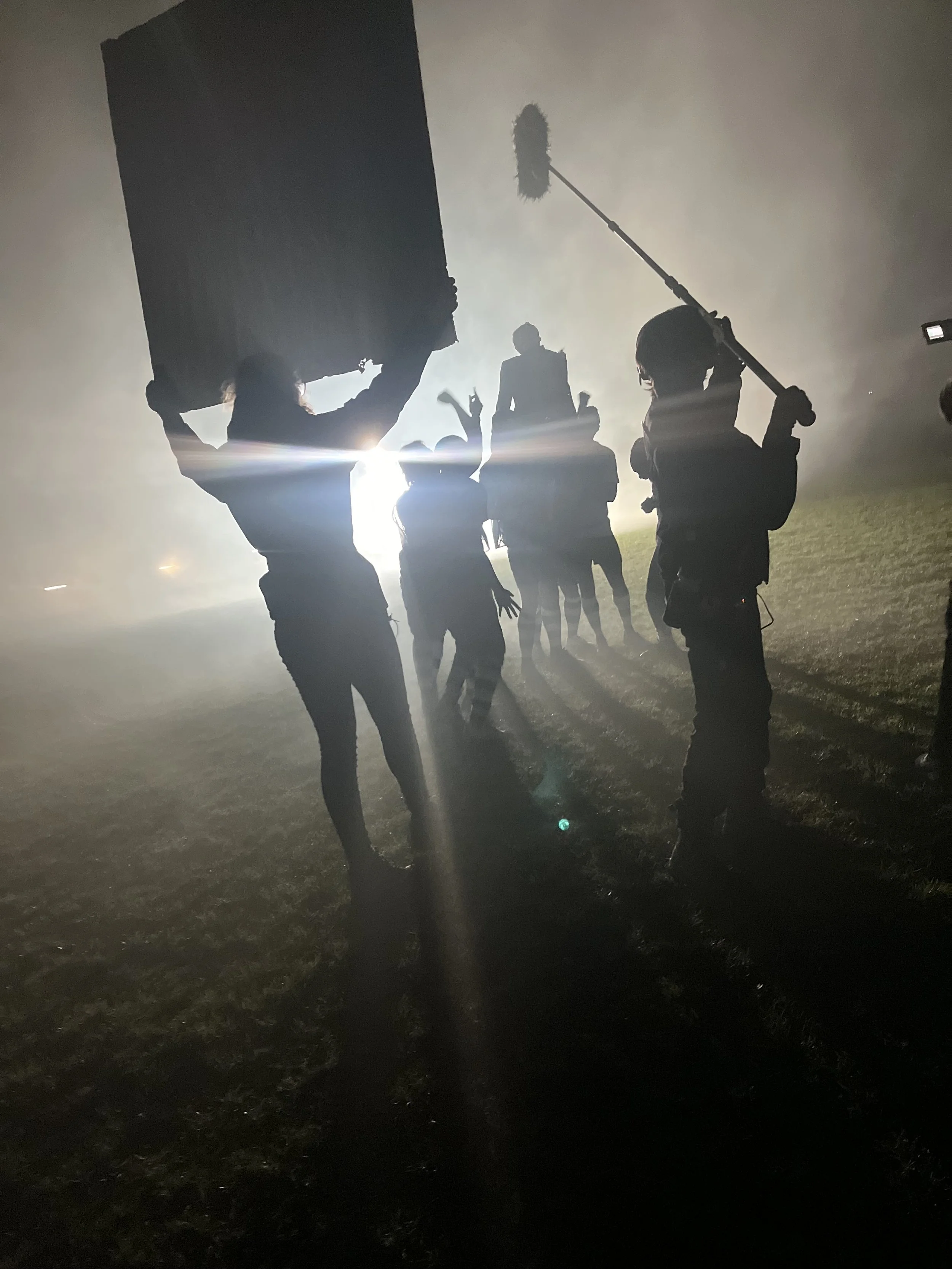 A film crew working at night, silhouetted against foggy back lights.