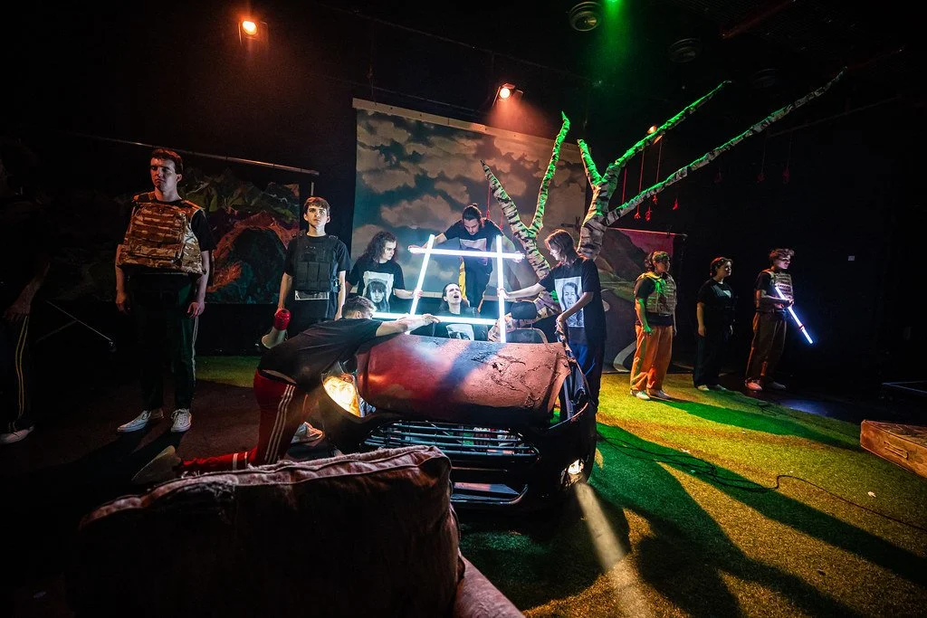 A theatrical stage scene with actors performing in front of a painted backdrop, featuring a leafless tree and landscape. The set includes a damaged red car, and some performers hold glowing props under dramatic lighting.