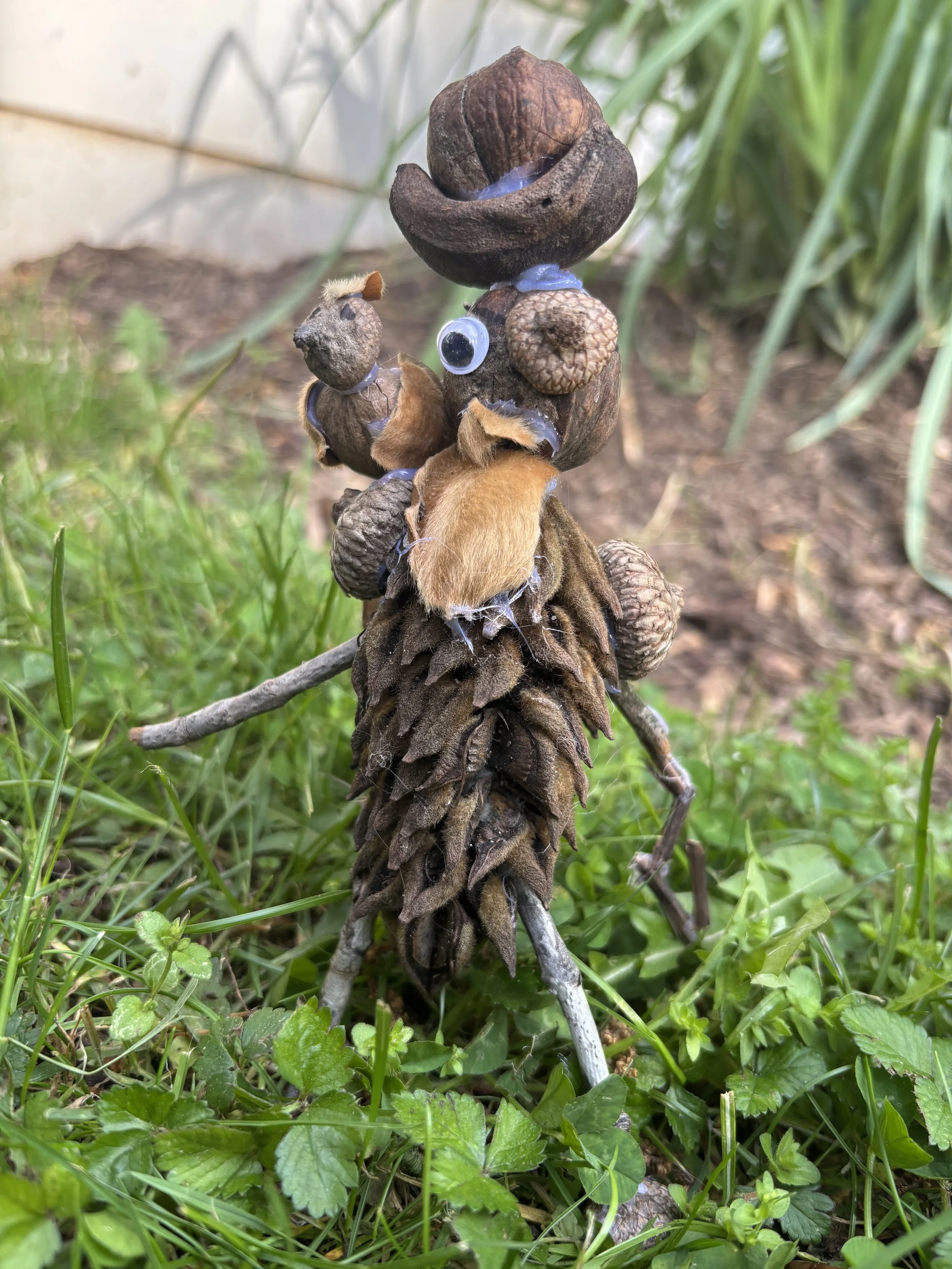 A pine cone with fake googly eyes and a small fuzzy toy attached, standing upright on a stick in a grassy area.