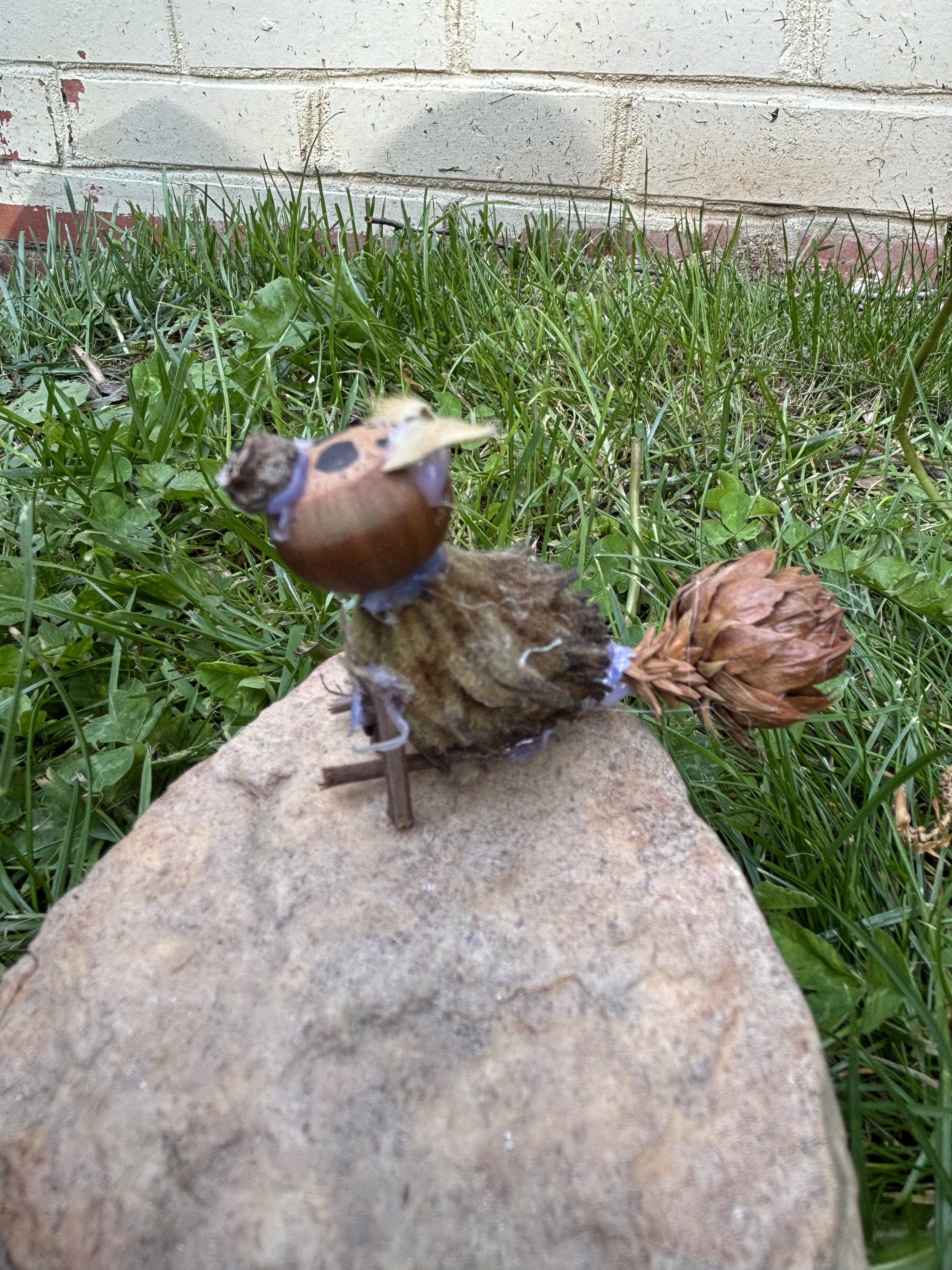 A dog like figure made from small pine cone glued to a miniature wooden stick, is placed on a stone surface with grass and a brick wall in the background.