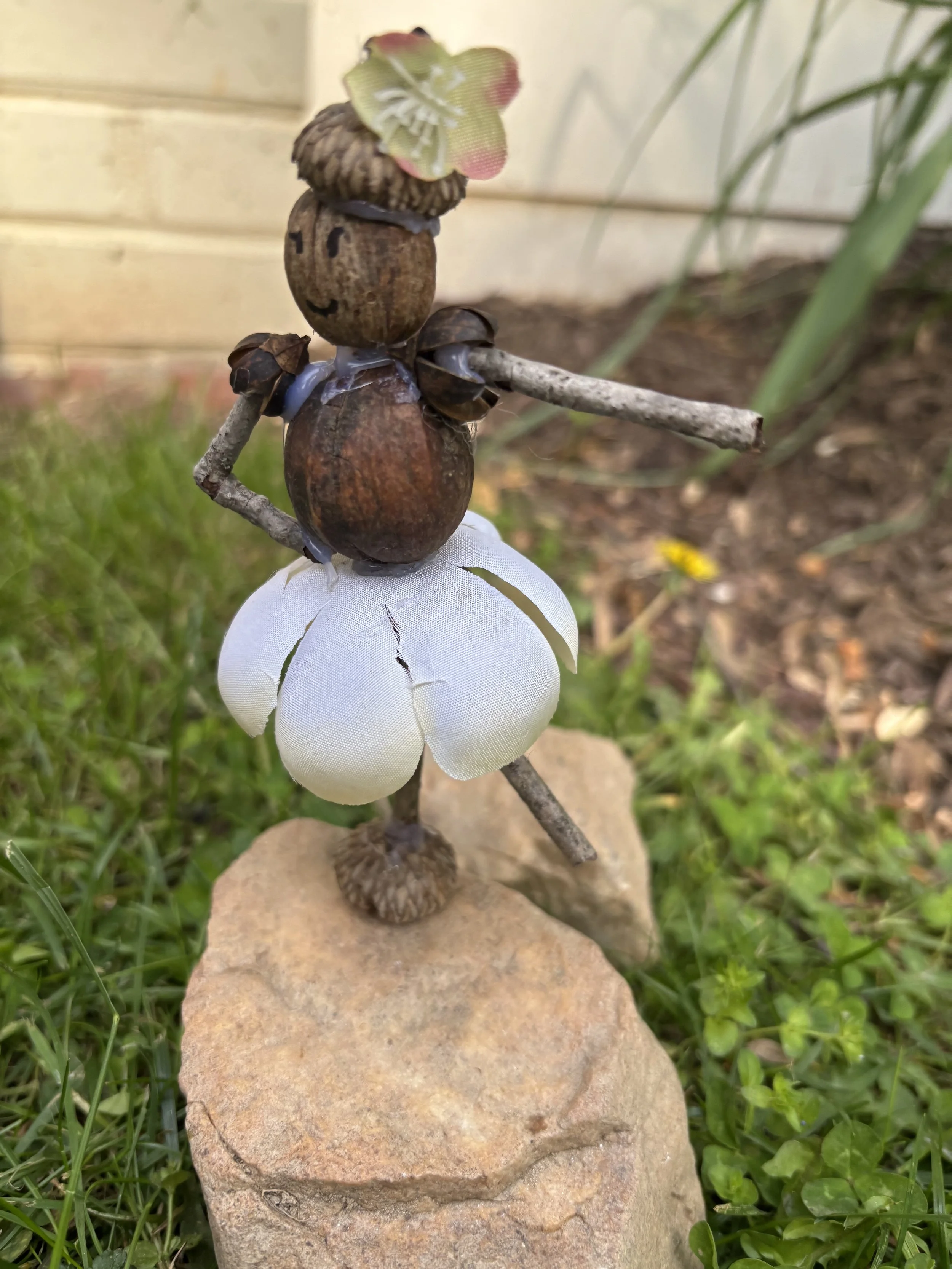 A small sculpture of a person sitting on a rock, made of natural materials like nuts, sticks, and leaves, with a flower on the head, set outdoors on grass.
