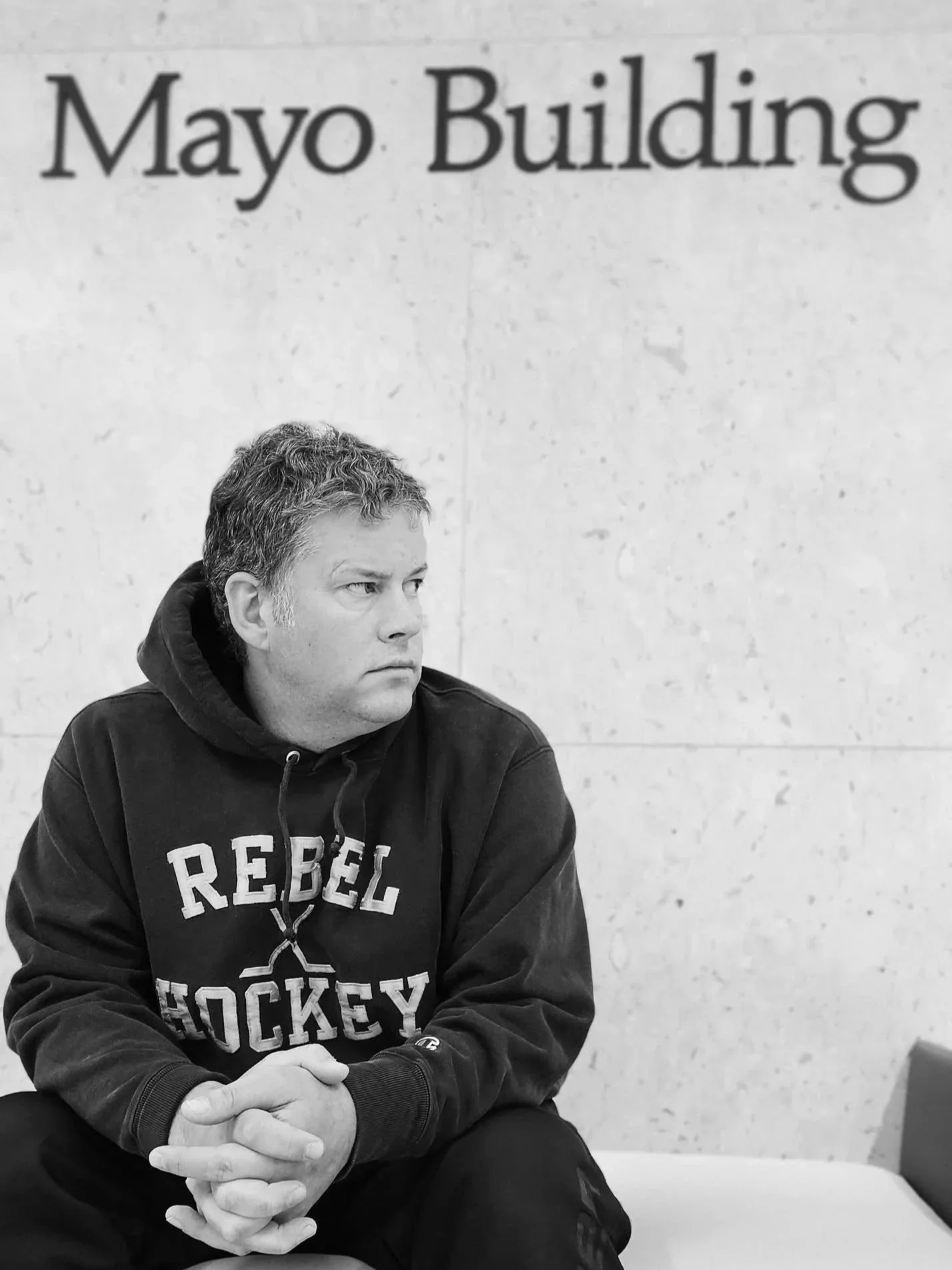 A man sitting indoors, wearing a hoodie that says 'Rebel Hockey'. He has curly hair and a serious expression, with his hands clasped together.