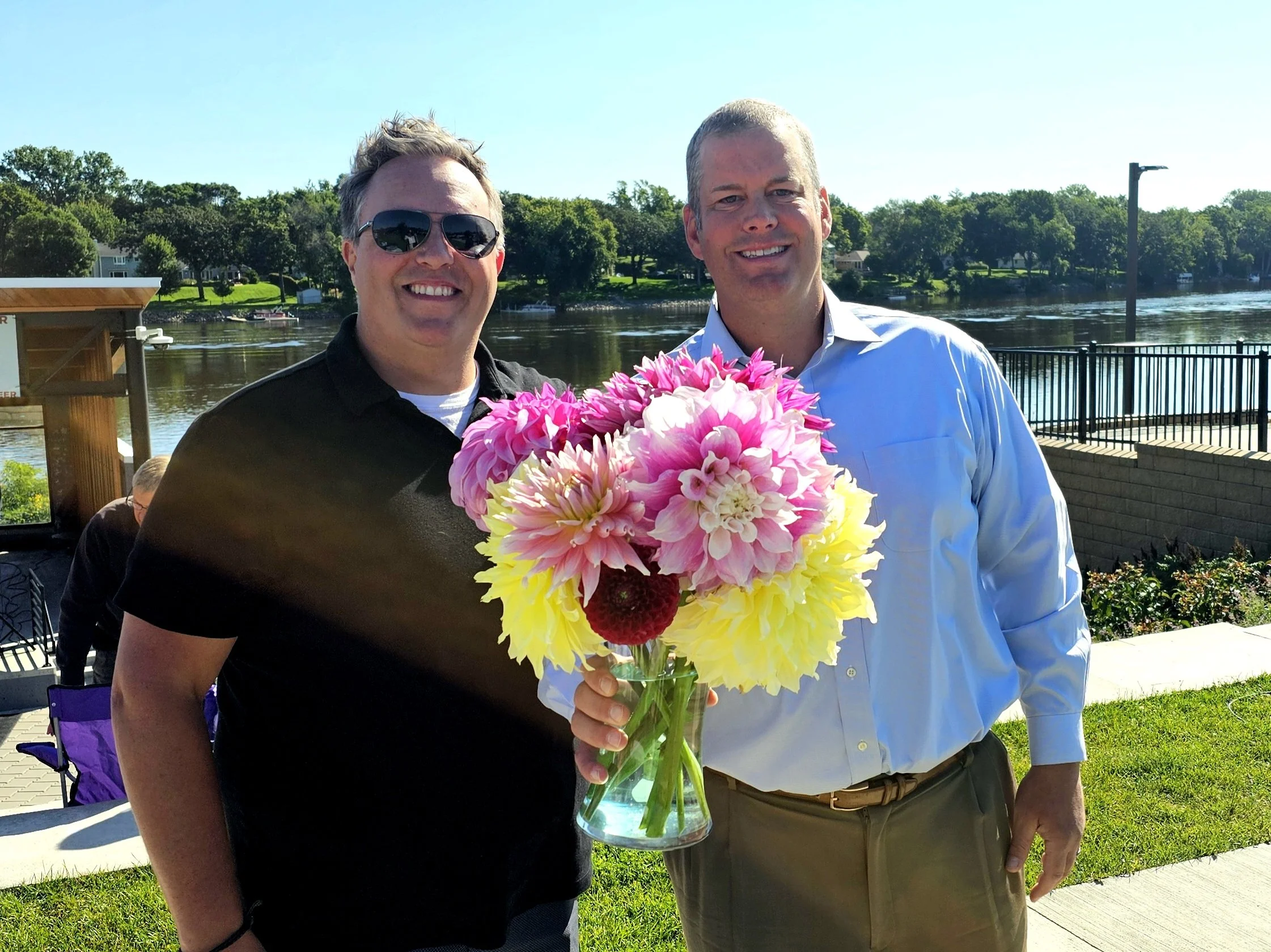 Two men standing together outdoors by a river; one man is holding a large bouquet of pink and yellow flowers in a glass vase. The man on the left is wearing sunglasses and a black shirt, and the man on the right is wearing a light blue shirt. The bac