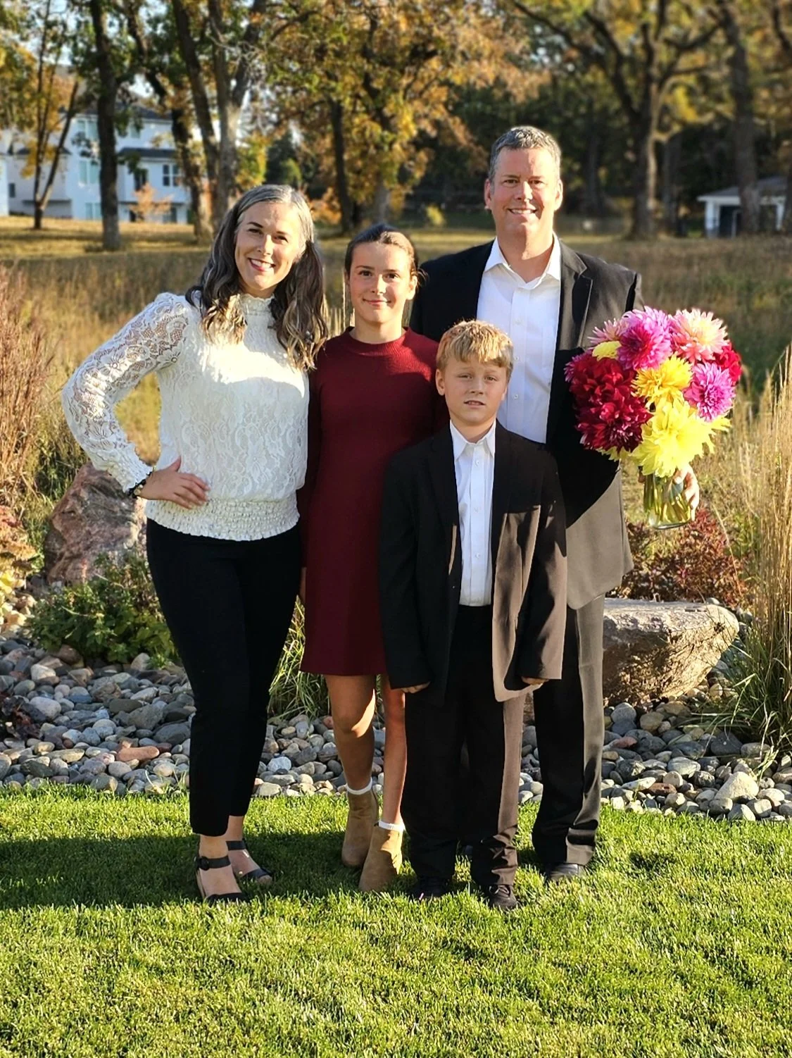 A family of five standing outdoors in a park with autumn trees in the background. The woman on the left is wearing a white lace sweater and black pants. Next to her is a girl in a red dress and beige boots. A young boy in a black suit and white shirt