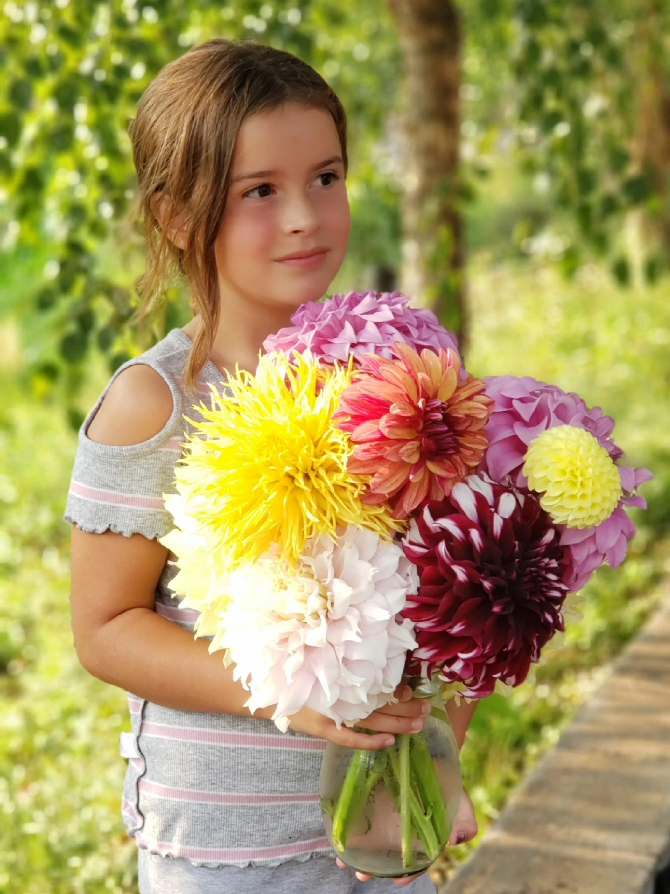 A young girl holding a large bouquet of colorful flowers outdoors with trees and a path in the background.