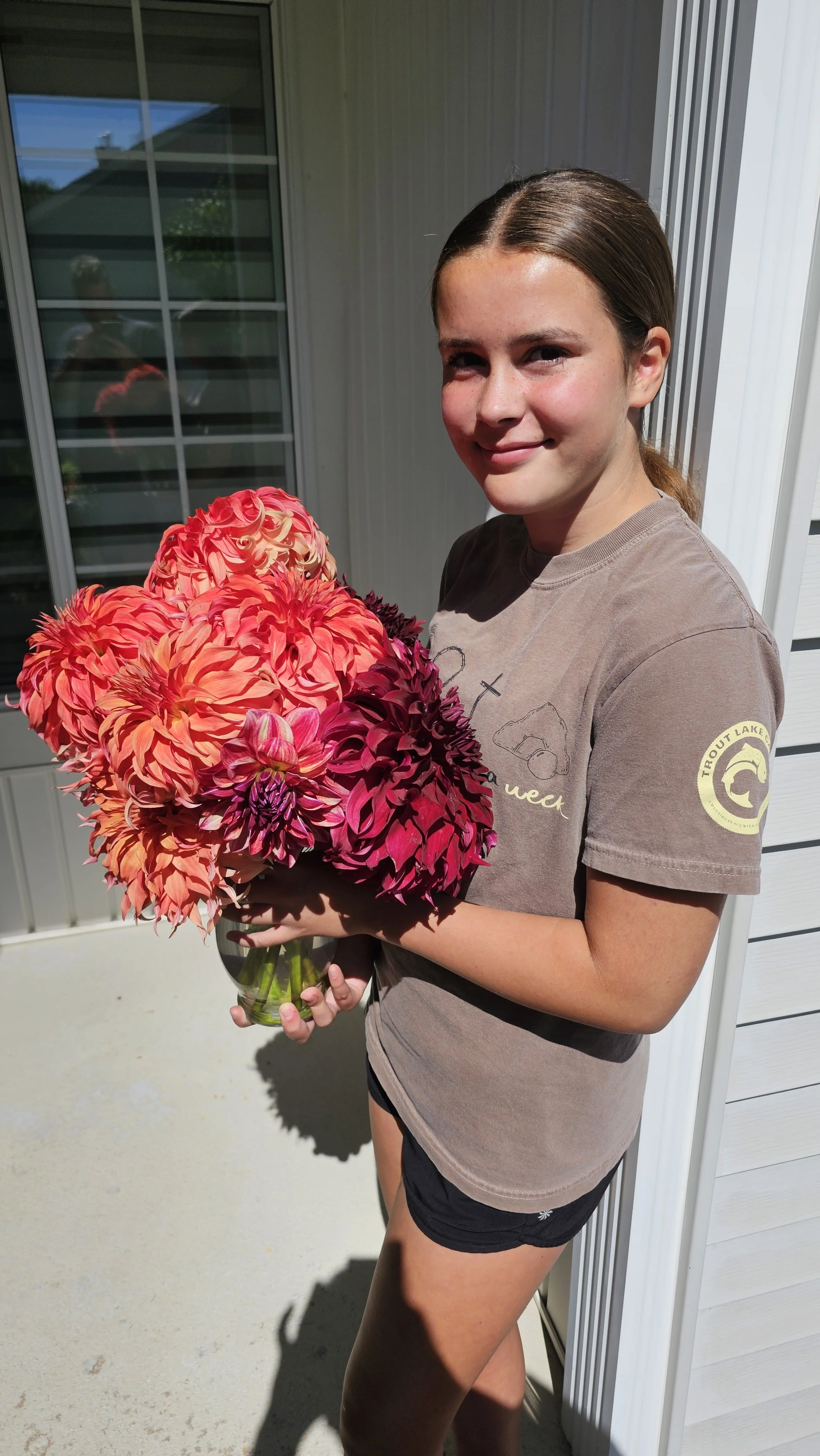 A girl with brown hair in a ponytail holding a large bouquet of bright pink, orange, and purple flowers standing outside on a porch.