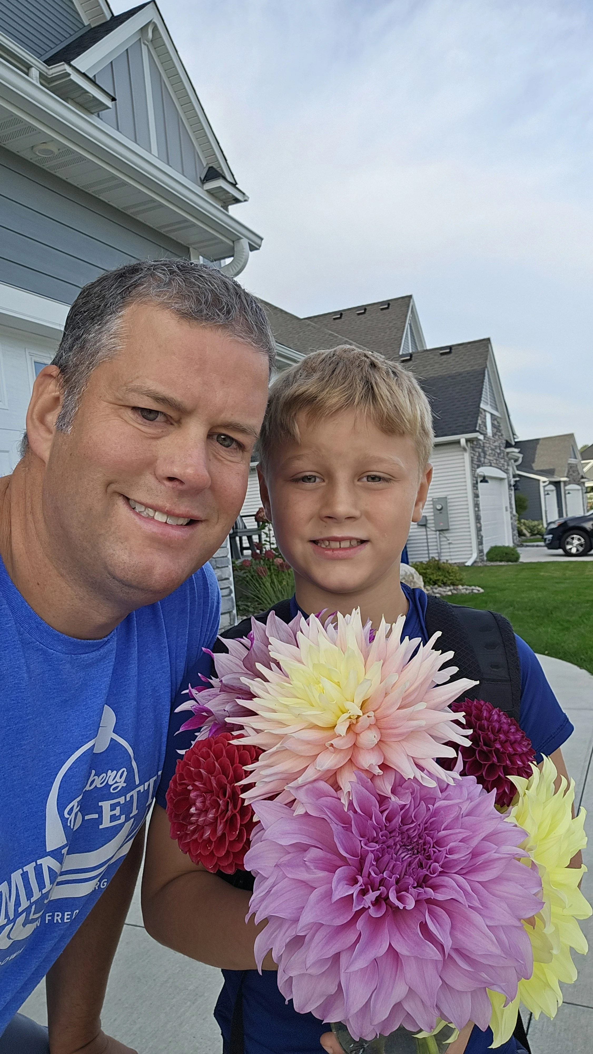 A man and a boy smiling and holding a bouquet of colorful dahlias outdoors in a suburban neighborhood with houses and cars in the background.