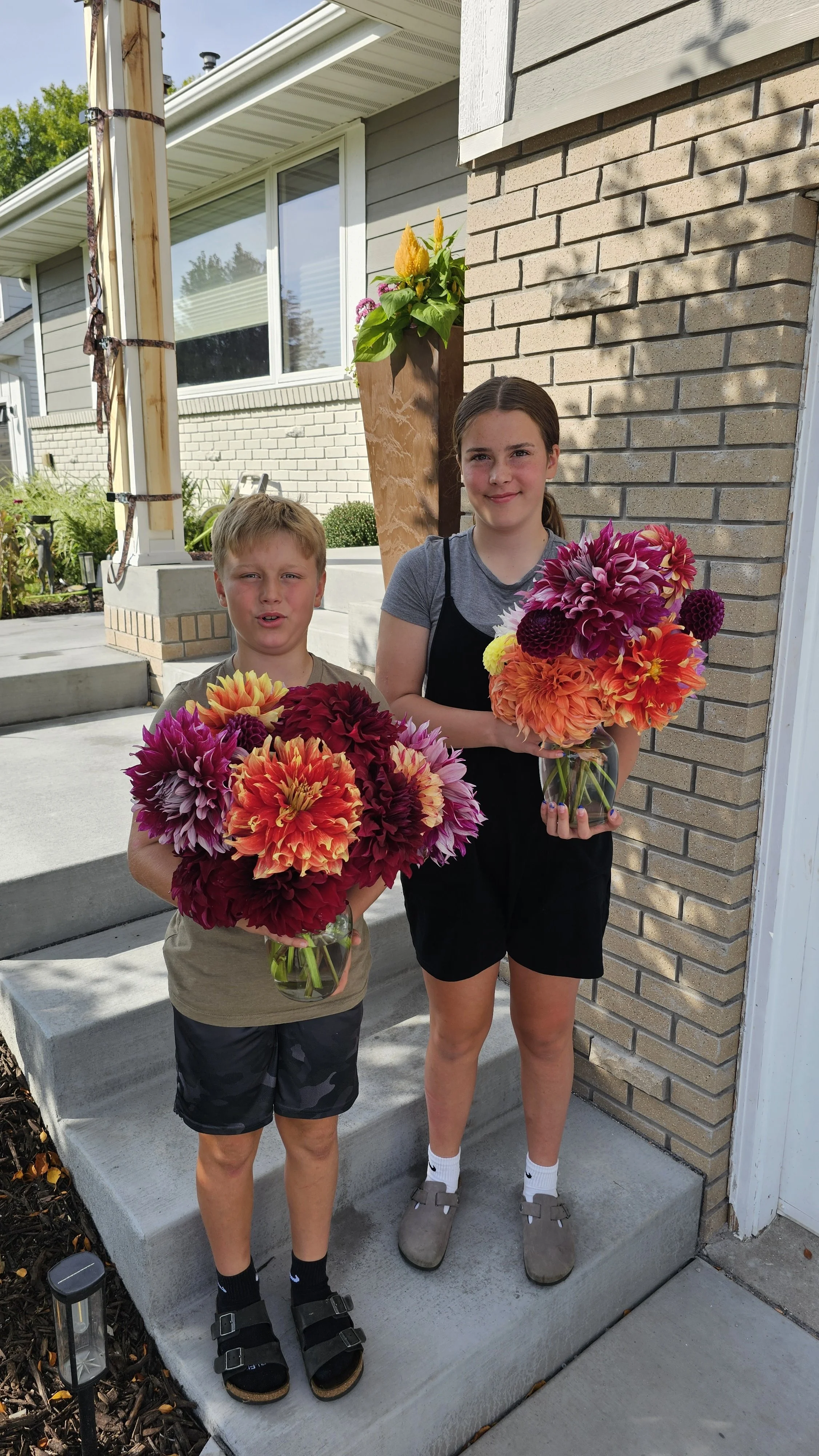 A young boy and a teenage girl standing outside on the steps of a house, each holding a bouquet of colorful flowers. The boy wears a beige t-shirt, camo shorts, and sandals with socks. The girl wears a gray T-shirt, black shorts, and gray sneakers, w