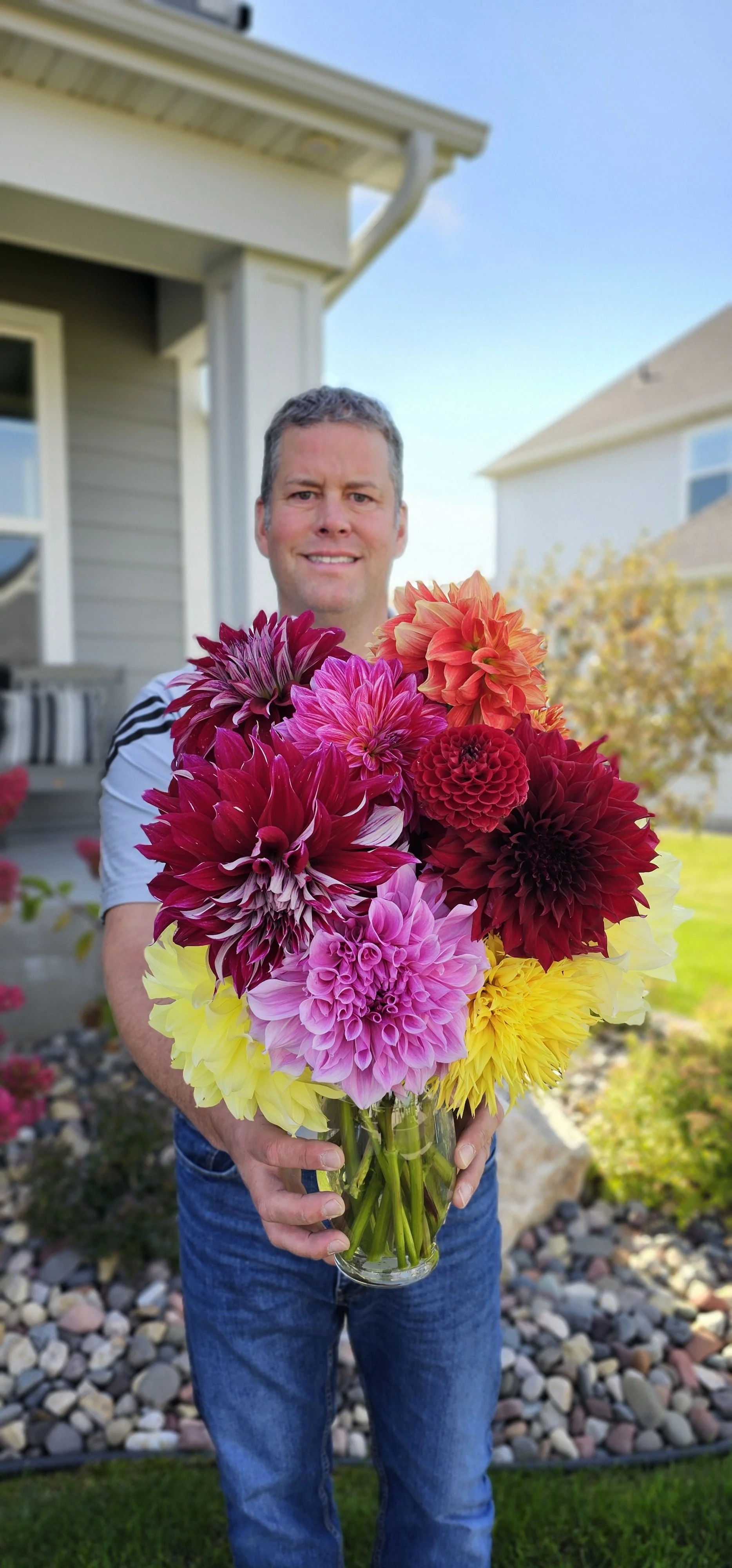 Man holding a large bouquet of colorful dahlias in front of a house with a garden.
