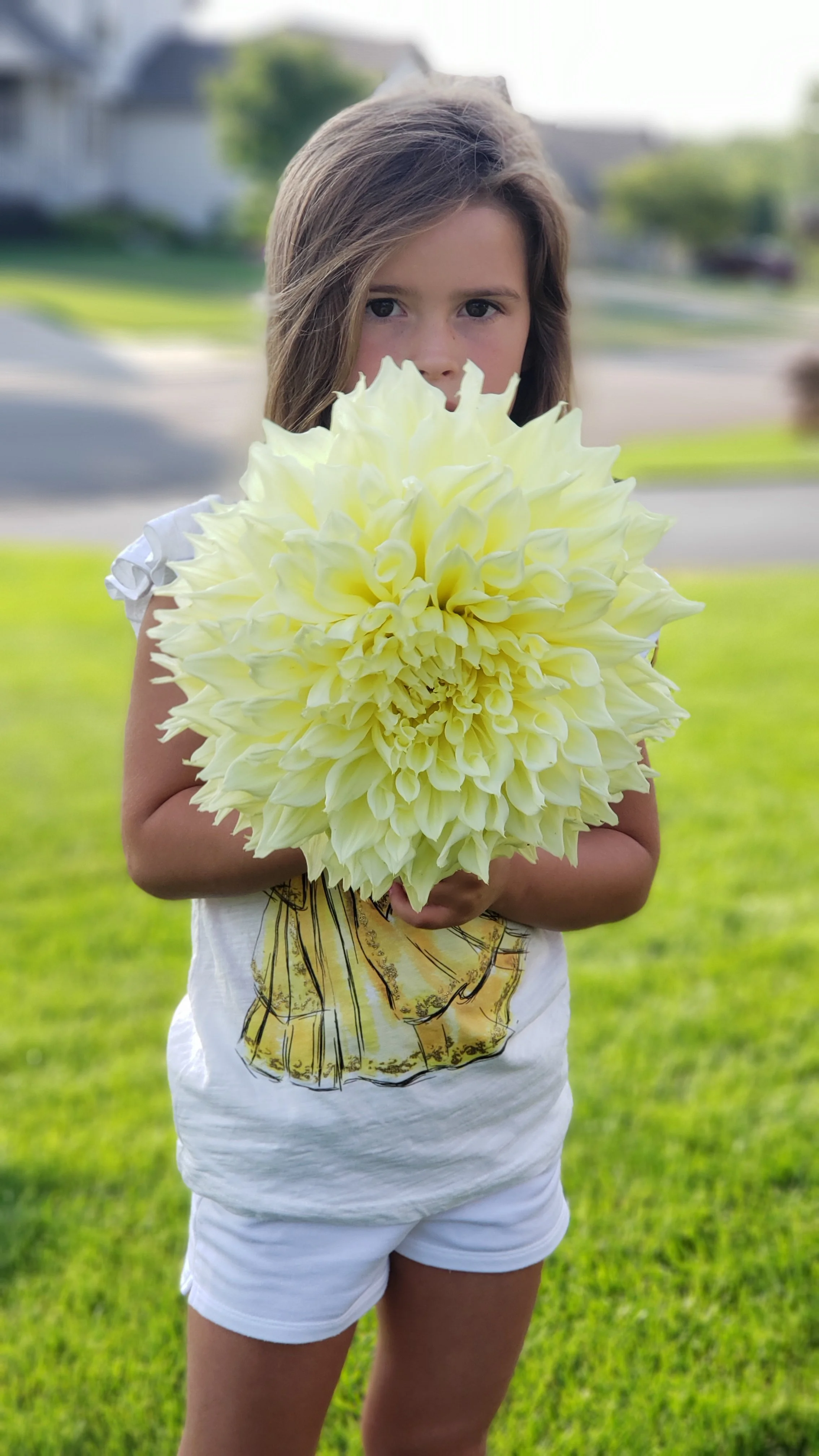 Young girl standing outdoors holding a large white and yellow flower in front of her face, with greenery and houses in the background.