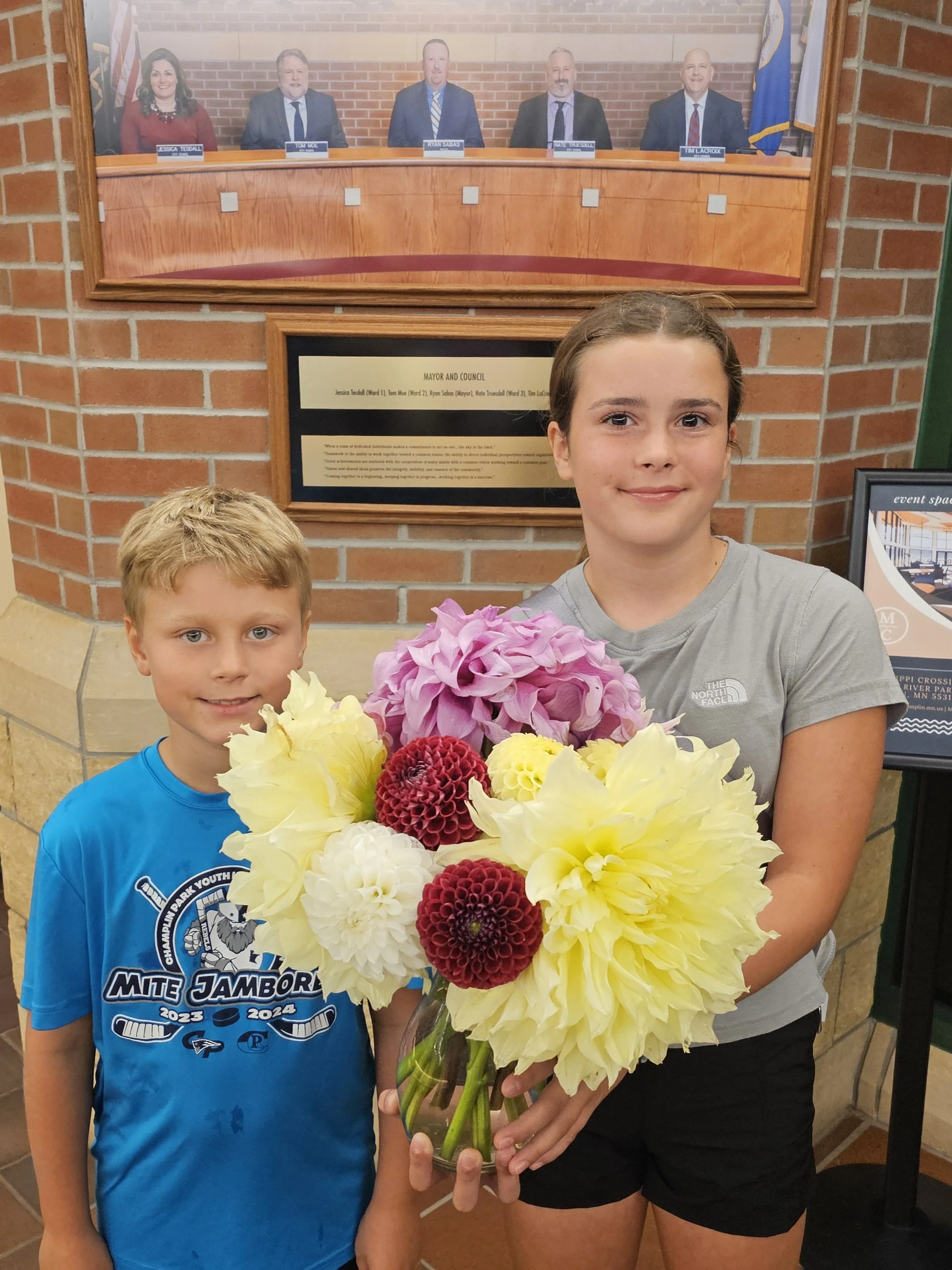 Two children, a boy and a girl, standing in front of a brick wall, holding a large bouquet of colorful flowers, with a framed photo of five people in formal attire displayed behind them.