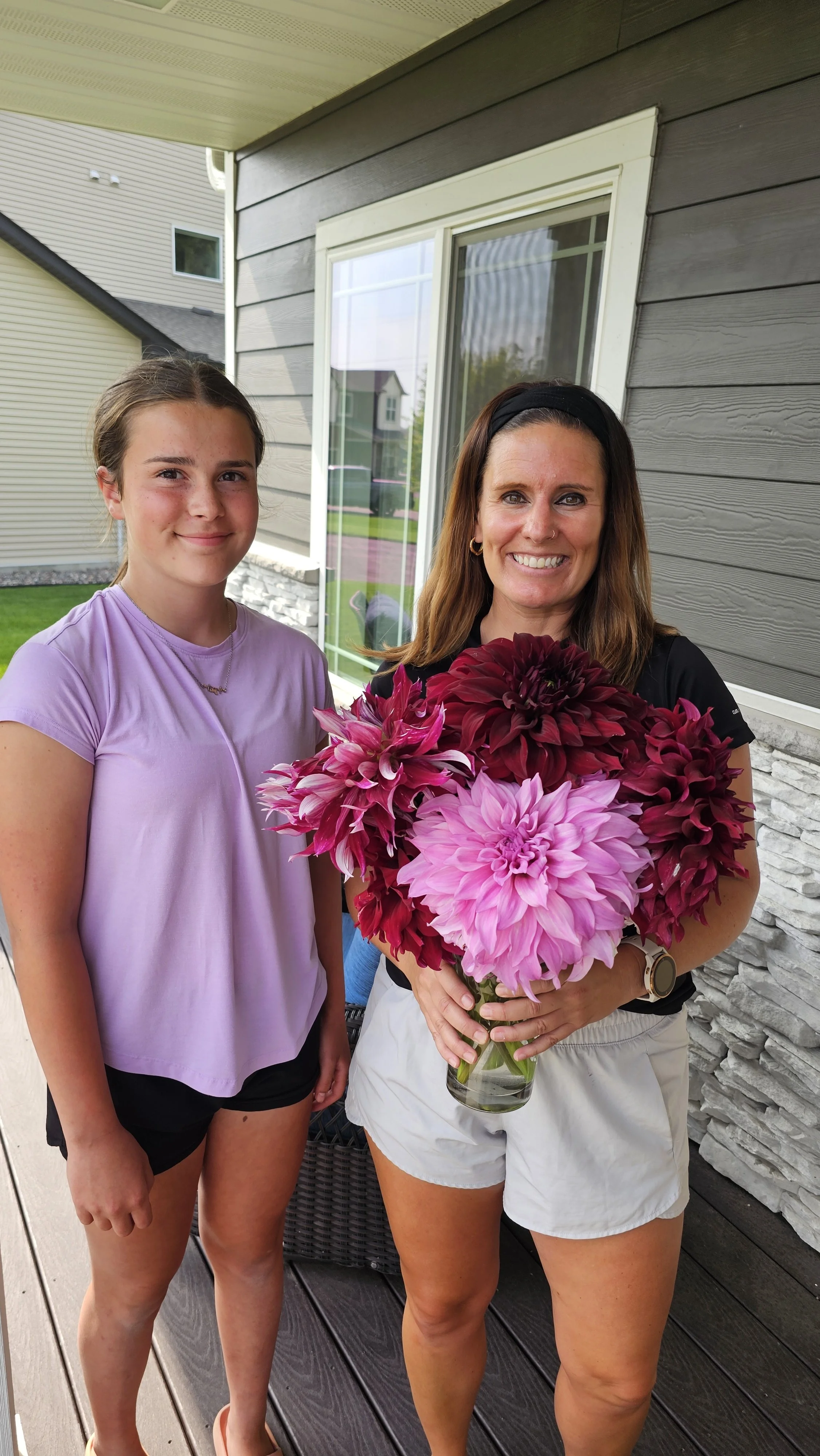 Two women standing on a porch, one holding a bouquet of large pink and dark red dahlias, both smiling at the camera.