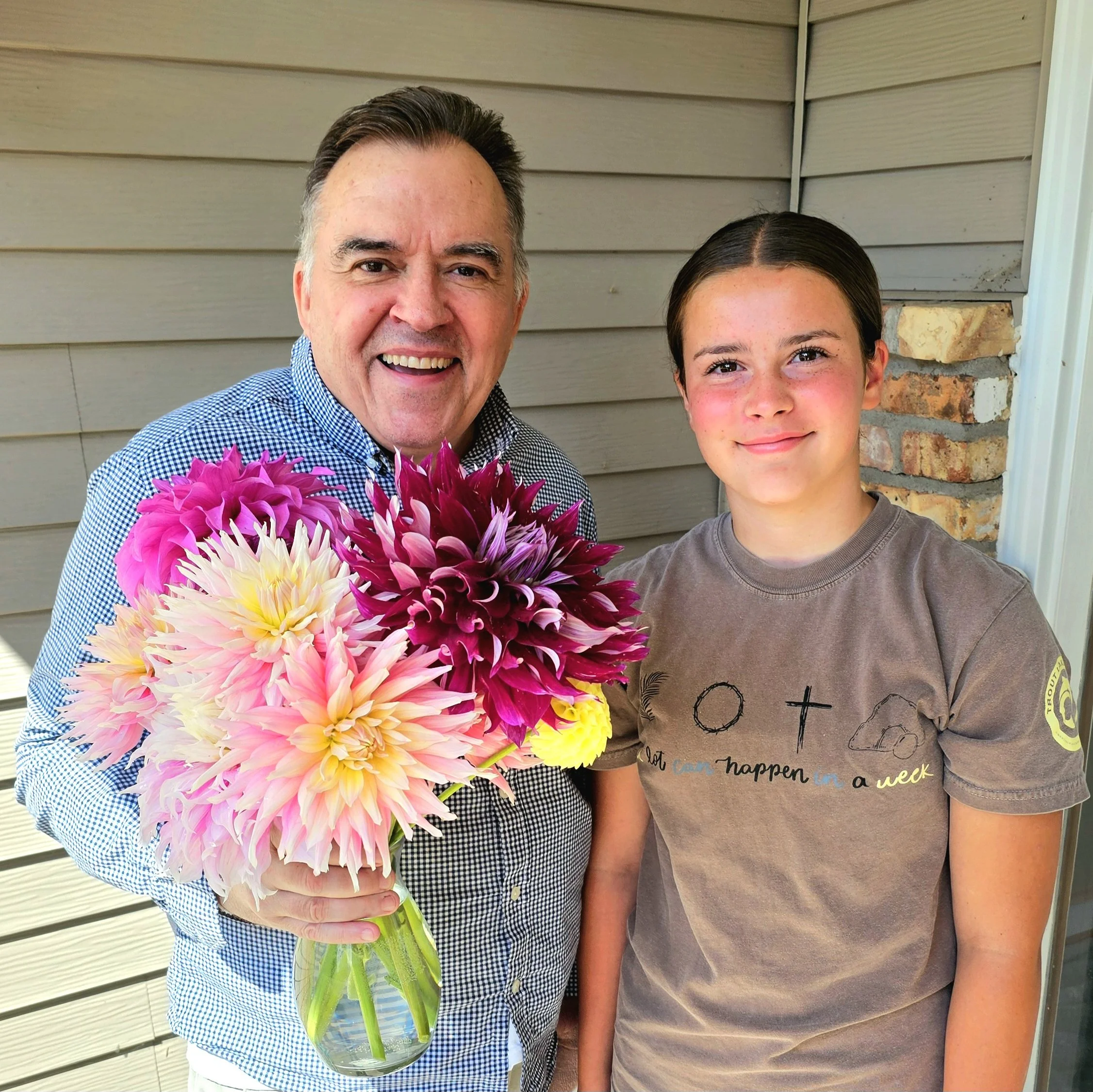 A man and a young girl holding a large bouquet of pink, purple, white, and yellow flowers in front of a house with beige siding and brick accents.