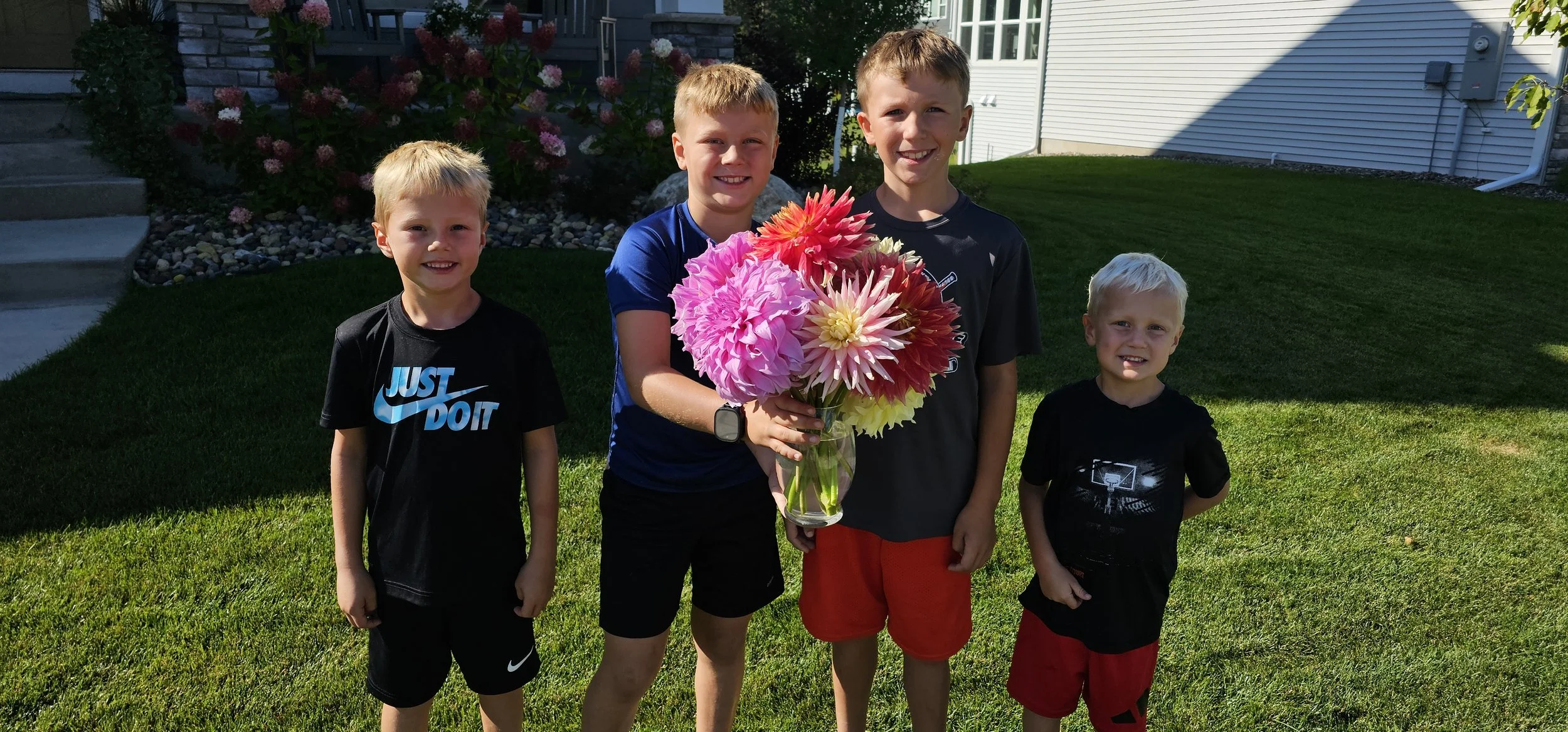 Four boys standing outside on grass, with flowers and a house in the background. The second boy from the left is holding a large bouquet of colorful flowers and smiling.